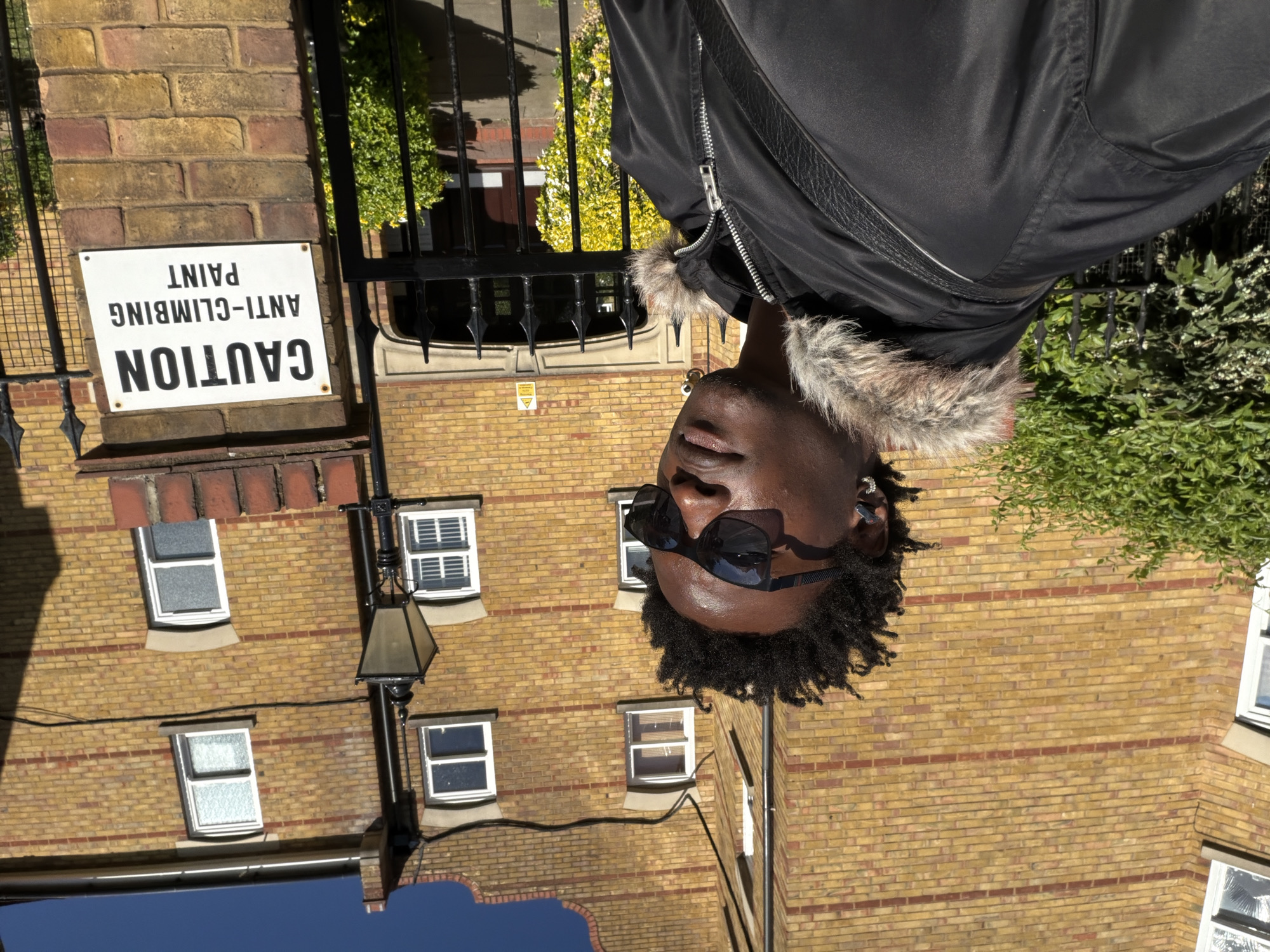 The image is a low-angle, vertical shot of a man in front of a brick building. The man is wearing sunglasses and a black jacket with fur trim around the collar. He appears to be facing the camera, his head tilted slightly. The building behind him is made of yellow bricks with red brick accents, and features multiple windows, a streetlight, and a sign that reads "CAUTION ANTI-CLIMBING PAINT". A black iron fence is partially visible behind the building. The image appears to be taken during the day. The location is likely in London, United Kingdom, based on architectural style and the sign.