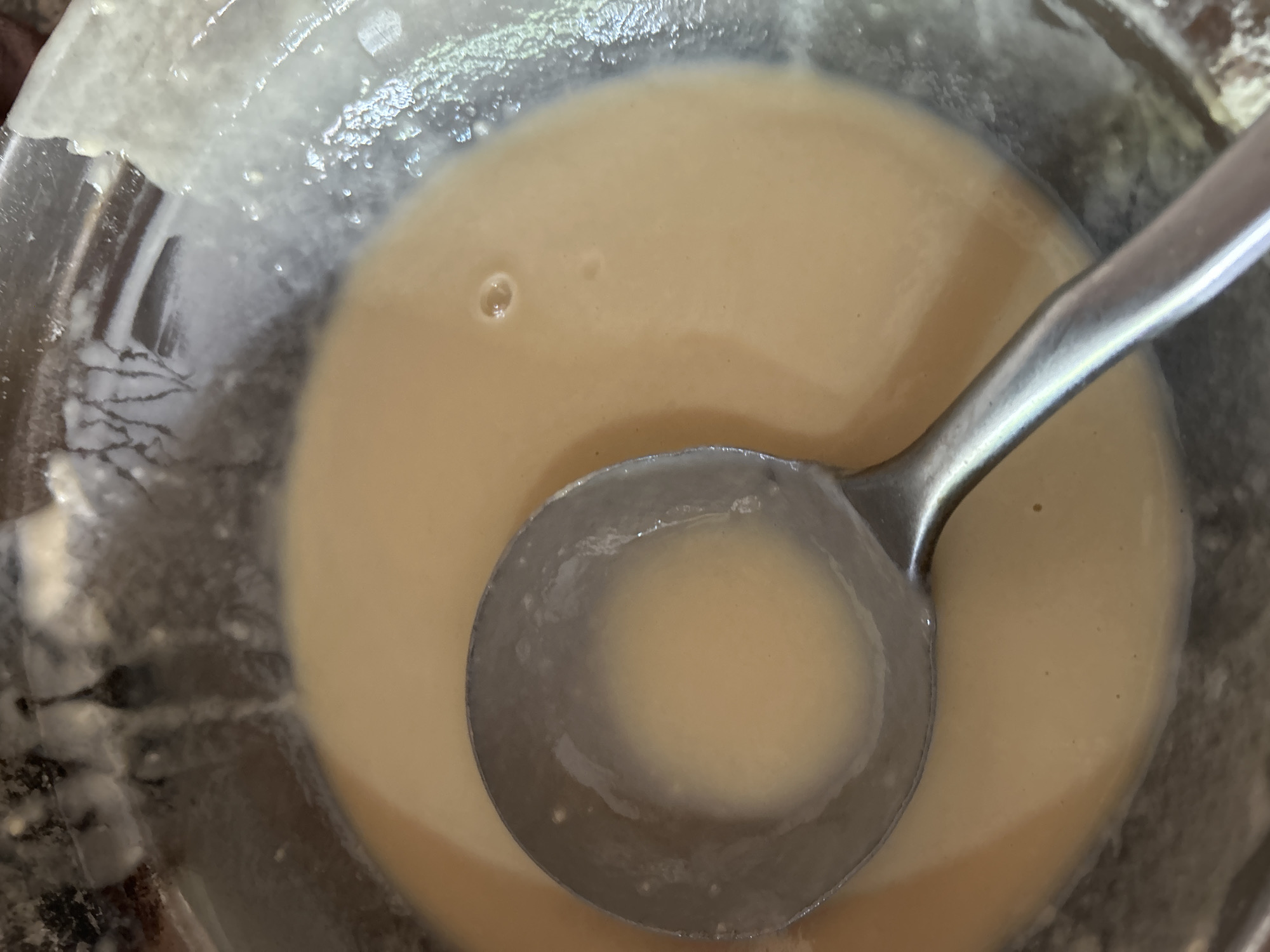 An overhead shot captures a close-up of a metal ladle filled with a light-colored, creamy liquid. The ladle rests within a larger, translucent bowl containing more of the same liquid. The bowl appears to be made of glass or a similar material and shows some residue along its inner rim. The liquid itself has a smooth, uniform consistency with a few small bubbles visible on its surface. The background is blurred but suggests a dark, possibly wooden surface. The scene is entirely indoors, likely in a domestic setting like a kitchen in Santa Ana, Venezuela.