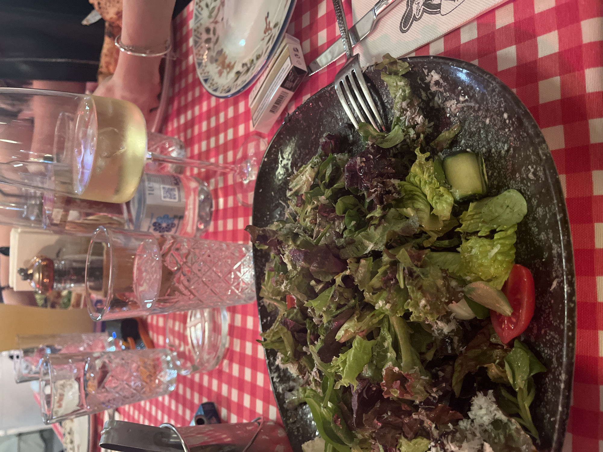 The image captures a dining table scene, viewed from an overhead perspective and rotated ninety degrees clockwise. The primary focus is a dark, irregularly shaped plate filled with a fresh green salad, comprising mixed lettuce (green and red varieties), cucumber slices, tomato wedges, and what appears to be grated Parmesan cheese. A silver fork rests on the edge of the salad plate.
Around the salad plate, the table is set for multiple diners. Visible objects include several clear drinking glasses of varying designs, including one champagne flute containing a pale golden liquid, likely white wine or champagne. A decorative, empty plate with blue and orange patterns is positioned behind the salad plate. Adjacent to it is a small white box, possibly a cigarette pack, with "B&H" clearly visible on its side, along with a partially visible QR code on a bottle label beneath one of the glasses. A knife with a textured handle and a napkin featuring a black and white illustration are also present. In the background, part of a person's arm and hand, adorned with a silver bracelet, are visible, reaching towards the table. Another hand is vaguely seen in the upper left corner.
The table is covered with a classic red and white checkered tablecloth, giving the setting a casual and possibly rustic or trattoria-like ambiance. A salt or pepper shaker can be seen in the distant background. The lighting suggests an indoor setting, likely a restaurant or cafe, possibly during the evening. The scene depicts people engaged in a meal or social gathering in Plovdiv, Bulgaria.