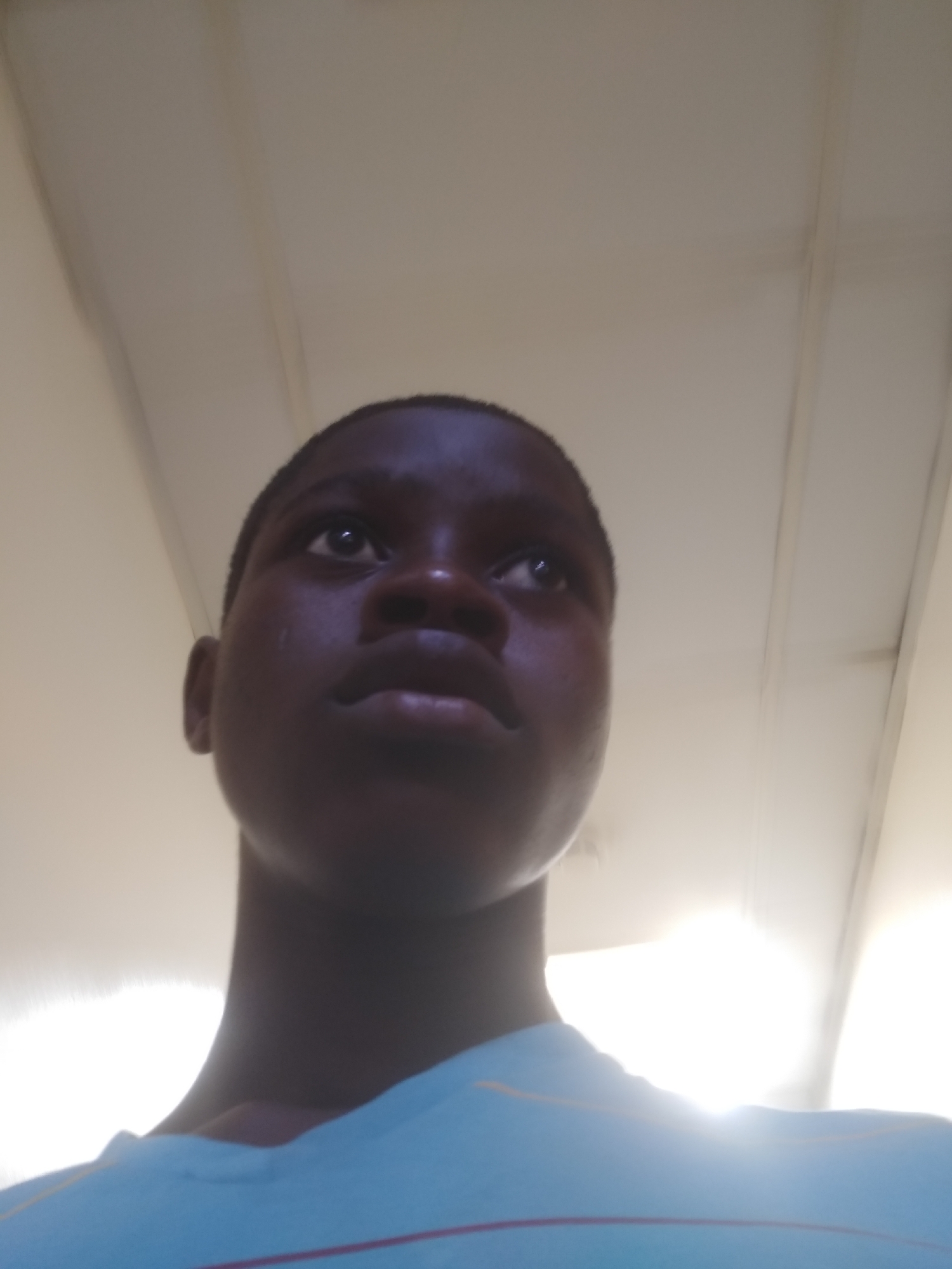 A low-angle shot shows a young dark-skinned person looking up. The person is wearing a light blue shirt with red and yellow stripes near the collar. The background consists of a blurry white ceiling with what appears to be light fixtures. Location cannot be determined.