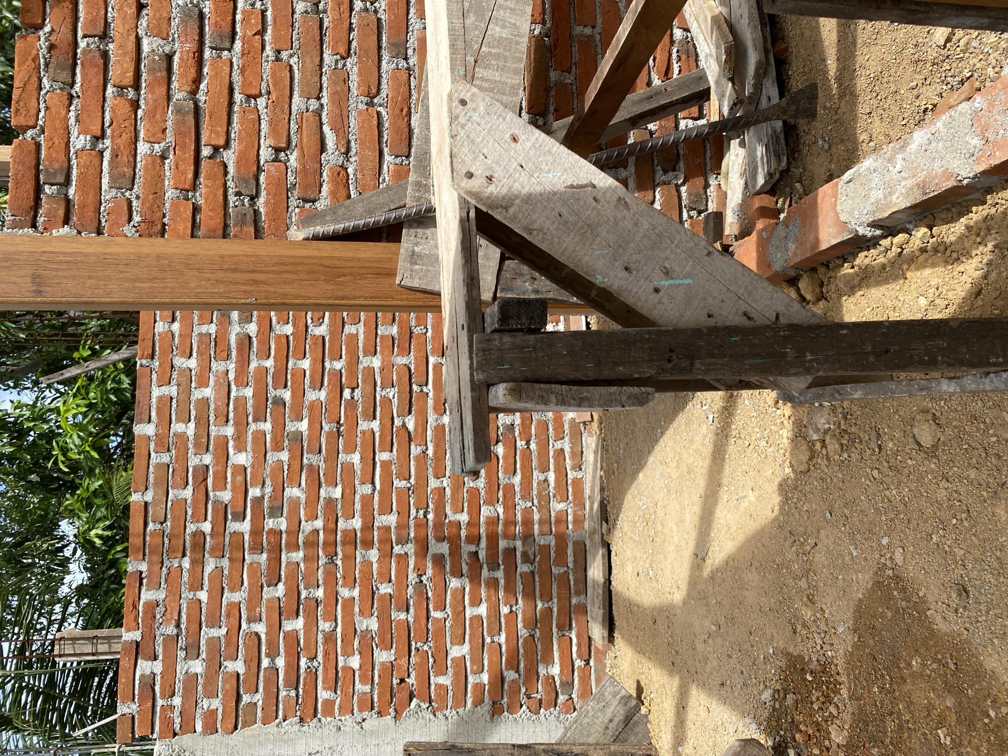 This is a close-up, outdoor, eye-level shot of a construction site. The foreground features a rough wooden scaffolding structure made of planks and supports. The ground is covered with loose, dry, sandy soil and small stones, with shadows cast from the scaffolding.
In the midground, a wall made of red bricks is visible, with some unfinished concrete on the left side. The bricks are laid in a standard running bond pattern with visible mortar. A large, vertical wooden post stands in front of the brick wall, partially obscuring it.
The background shows more of the brick wall and lush green foliage from trees, with a hint of blue sky visible above. The overall impression is a scene of ongoing construction in a somewhat rural or undeveloped area. There are no people or animals present in the image.