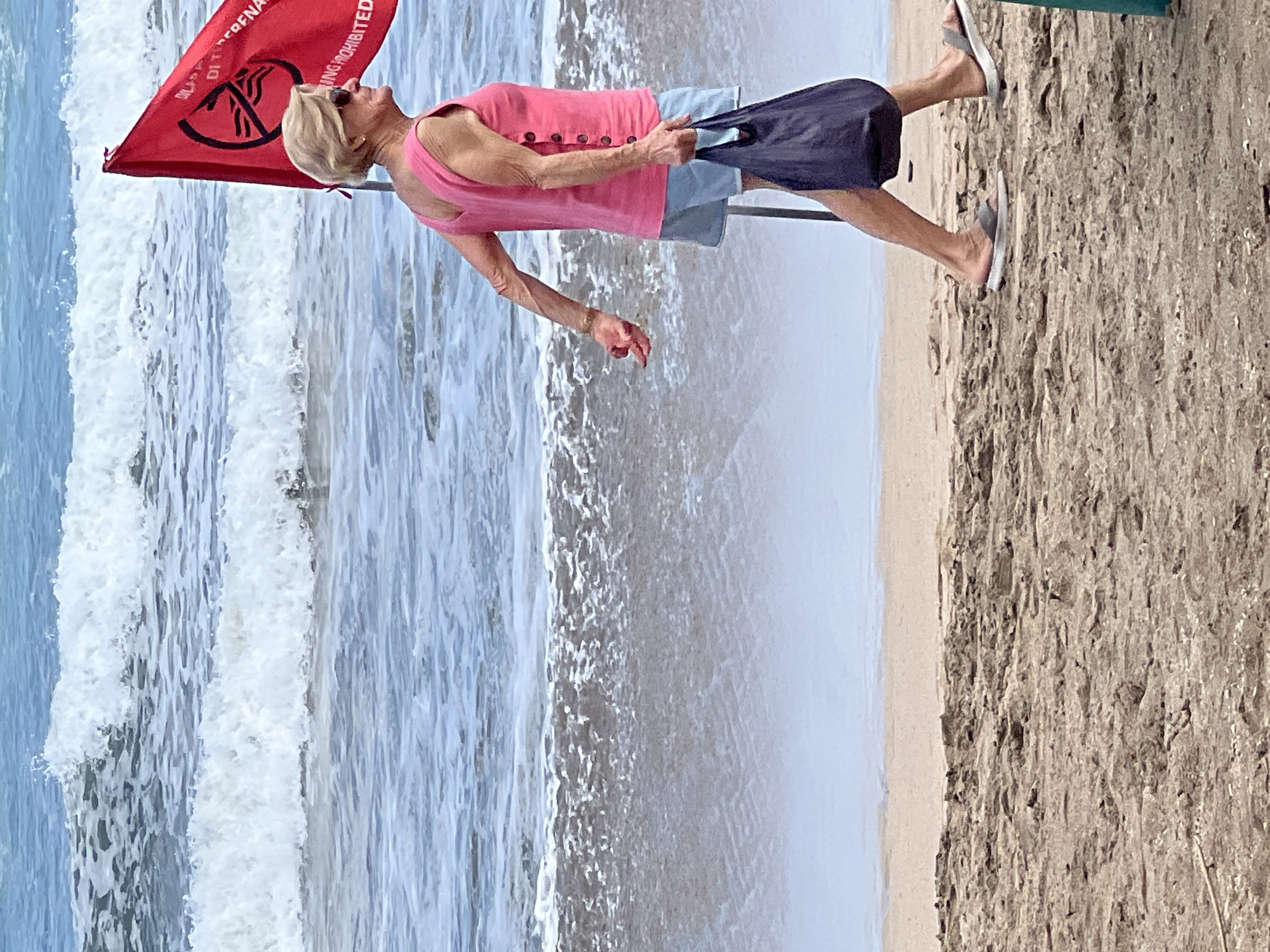 In Kuta, Indonesia, an older woman with blonde hair, wearing a pink sleeveless top and light blue shorts, walks along the sandy beach. She is carrying a dark blue tote bag in her right hand and wears sunglasses. A red flag with a symbol of a person swimming within a circle and a line through it, indicating swimming is prohibited, is planted in the sand to her left. Behind her, waves crash onto the shore under a clear sky. The background shows the ocean and a sandy beach.