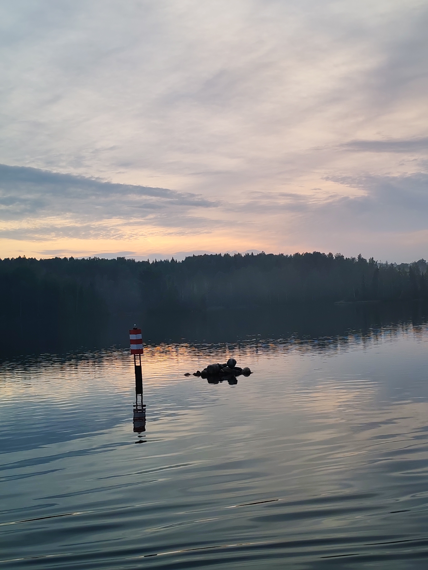The image depicts a serene lake scene at dawn or dusk, with the sky displaying soft pastel hues from sunrise or sunset. A navigational buoy, painted with alternating red and white bands, stands vertically in the water, casting a reflection. A small cluster of rocks is visible near the buoy. The background features a dense, dark forest lining the distant shore, with a light mist or fog settling over the trees. The water's surface is calm, showing ripples that reflect the muted colors of the sky and the surrounding environment. The location is identified as Kurikaevo, Russia.