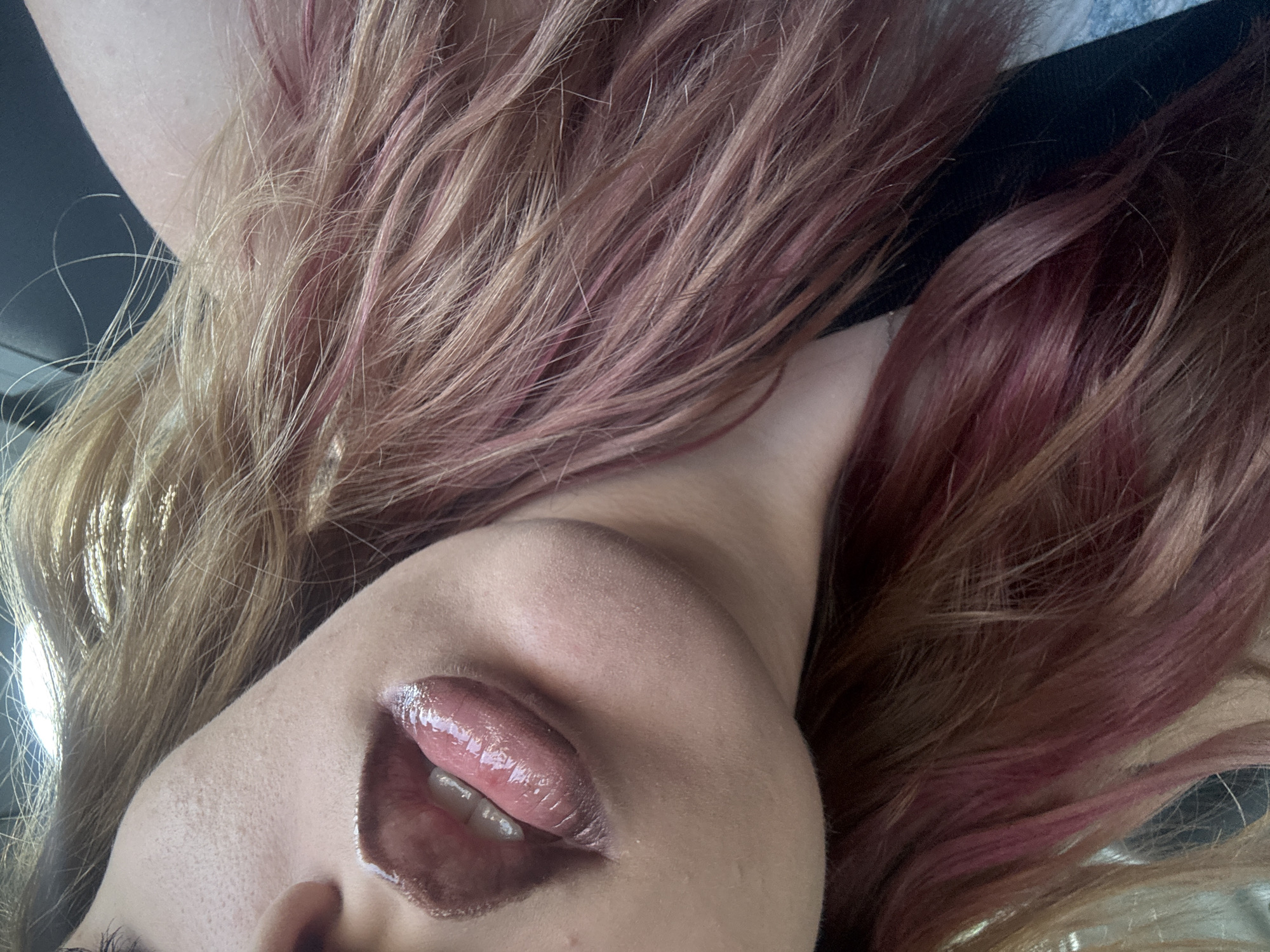 The image is a close-up, waist-up shot of a person's face, focusing on their mouth and hair. The person has full lips, glossed with a metallic, brownish-pink lipstick. Their tongue is visible, slightly protruding. The hair is a blend of blonde and pink, styled in loose waves. The background is blurred, suggesting the photo was taken indoors or in a moving vehicle. There is no indication of the location being Vratsa, Bulgaria, or any other specific city. The scene is a portrait of the individual's facial features and hair.