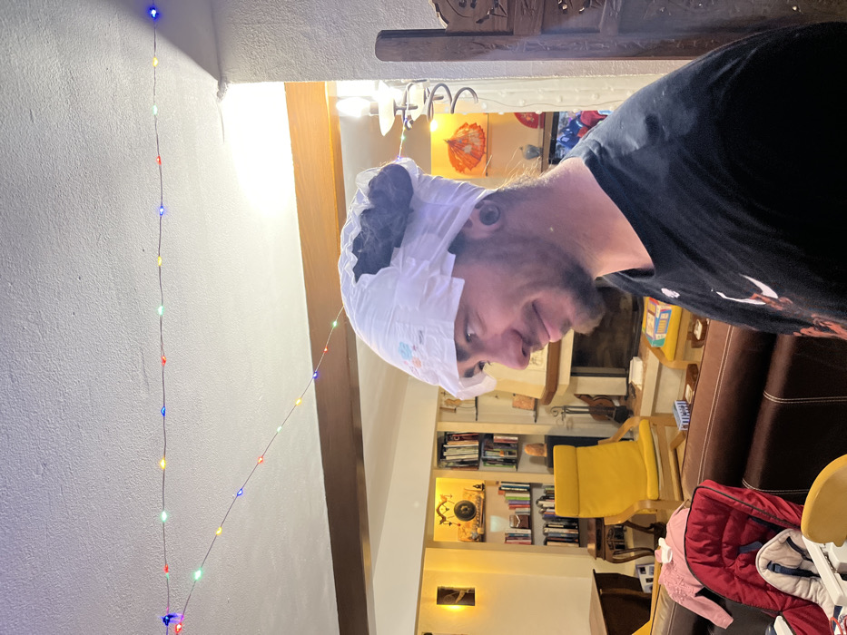 A person in a black t-shirt wears a diaper on their head indoors. String lights hang near a wall and doorway. A living room is visible in the background with a bookshelf, yellow chair, and brown couch. A jacket is partially visible near the bottom of the frame.