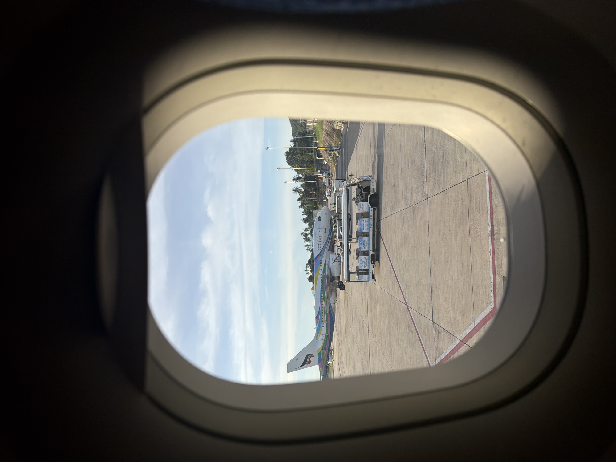 The image, taken from an airplane window, shows an airport tarmac. An airplane with colorful livery, possibly belonging to Bangkok Airways, is parked on the tarmac. In front of the airplane, a baggage cart is visible with personnel attending to it. The scene is set against a backdrop of a bright, cloudy sky and a line of palm trees, suggesting a tropical location. The airport markings on the tarmac are clearly visible. This scene is likely at the Samui International Airport, located in Baan Bang Rak, Thailand.