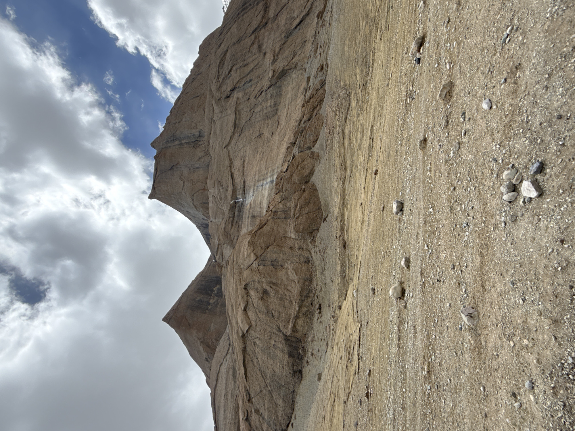 The image shows a towering rock formation in a rugged landscape under a cloudy sky with patches of blue. The rock face is primarily tan and brown, marked with textured striations and crevices. A dirt and gravel-covered ground extends from the base of the rock formation into the foreground, scattered with various-sized stones. There are no people visible in the scene. The location is unspecified, though it's claimed to be in China, Parga town.