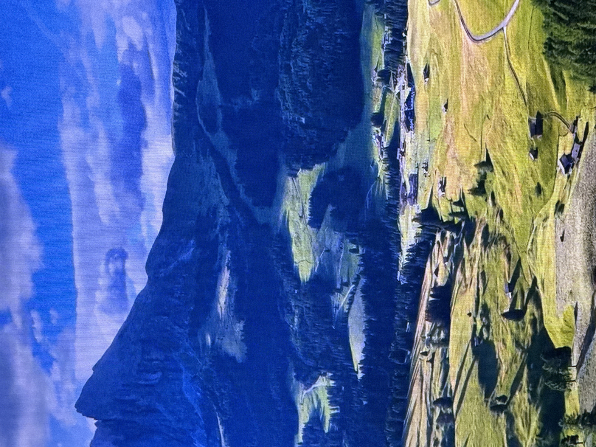 This is an aerial, high-angle landscape photograph of a mountainous region with scattered farmhouses and dense forests. The scene is characterized by rolling green hills bathed in sunlight, with dark, shadowed valleys and a prominent, jagged mountain peak in the background. A winding road is visible in the lower right section of the image, leading through the terrain. The sky above is a vibrant blue, dotted with white and gray clouds. The image does not depict any urban environments, people, or activities. It is a natural landscape and therefore not located in Dubai, United Arab Emirates.