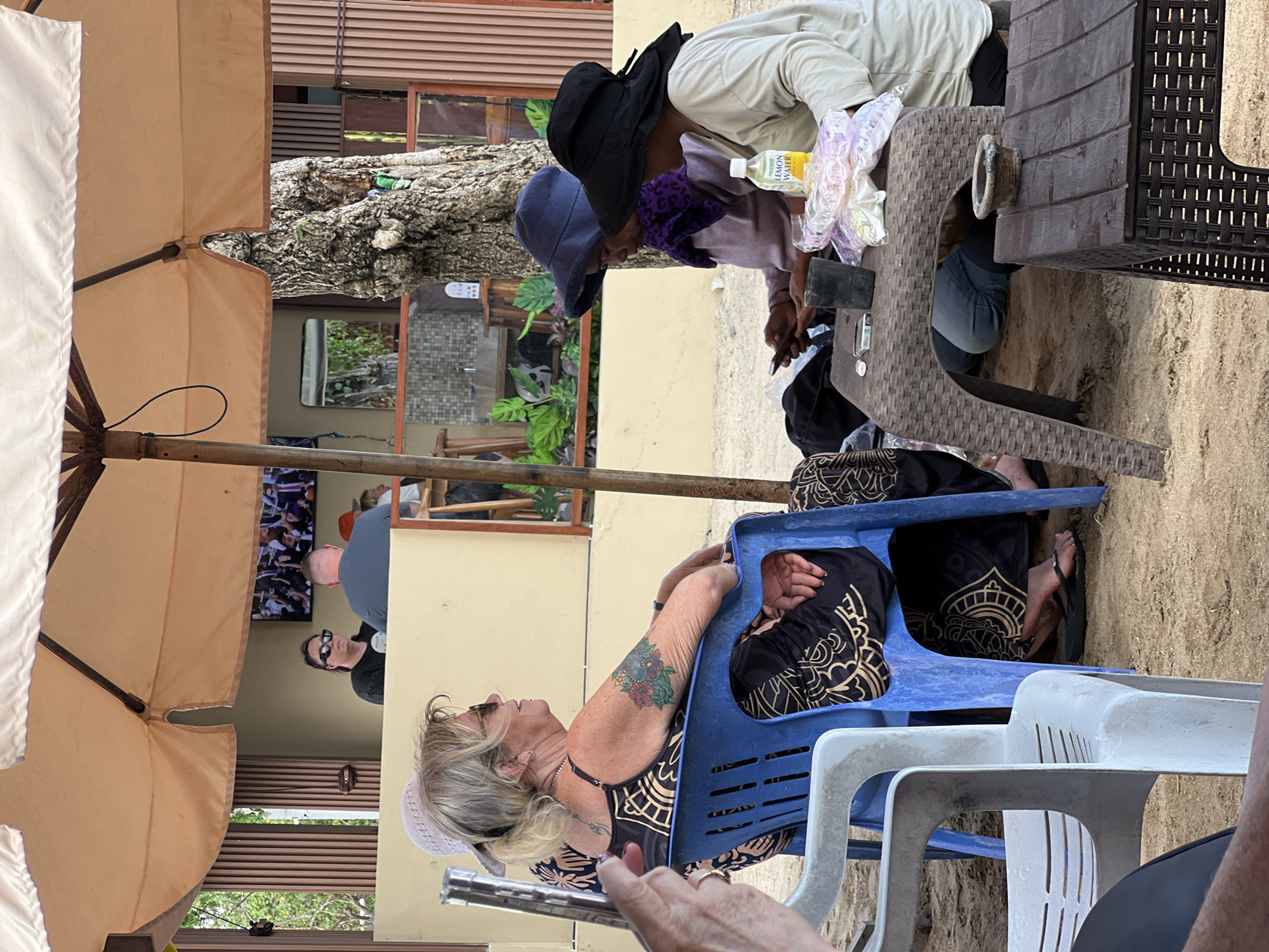 The image depicts a group of people seated at tables under a large umbrella, likely at a beachside cafe or restaurant in Kuta, Indonesia. Sand covers the ground. In the foreground, a person's arm is visible, suggesting the photographer is part of the scene.
Central to the image, a woman with blonde hair and sunglasses is seated in a blue plastic chair, facing right. She is wearing a black sleeveless dress with a gold and black pattern. Her left arm features a colorful floral tattoo. To her right, another person is partially visible in a similar dress.
Further back, two individuals are seated at a woven plastic table. The person on the left, wearing a purple scarf, is looking down at a mobile phone. The person on the right, wearing a wide-brimmed black hat and a light-colored long-sleeved shirt, also appears to be engaged with the phone. On the table between them are plastic-wrapped items, a bottle labeled "LEMON WATER," a dark cup, and a lighter. A second, lower brown table with a woven base is in the lower right, holding a small stone mortar.
In the background, behind the seated group, is a raised structure with a television displaying a sports event. Other patrons and a bar area are visible within this structure. The environment suggests a casual, outdoor gathering place, typical of Kuta's beachside establishments.