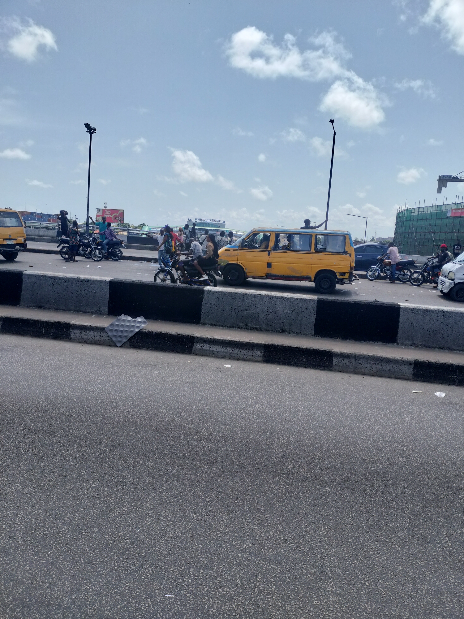 The image captures a vibrant daytime street scene in an urban environment, likely Lagos, Nigeria, given the distinctive yellow commercial van and numerous motorcycles. The perspective is from a lower vantage point, looking across an asphalt road and a concrete median barrier towards an elevated road or overpass where most of the activity is concentrated.
The sky is bright with scattered white clouds, suggesting a clear day. On the elevated section, a yellow commercial passenger van, commonly known as a "Danfo" in Lagos, is prominently featured, appearing to be stopped or moving slowly. Several people are visible in and around the van. Numerous motorcycles, known as "Okada" taxis, are also present, with riders and passengers. Some individuals are standing on the roadside or near the barrier, possibly waiting for transport or engaged in other activities. One person is seen with their arm raised, leaning on the back of a motorcycle.
To the left, another yellow vehicle, possibly another van, is partially visible. To the right, a white van and a dark car are also present. In the foreground, on the concrete barrier, a crumpled piece of metallic or plastic debris with a dimpled pattern lies. The main road surface in the immediate foreground appears empty.
In the background, overhead structures include lampposts. On the elevated section, various billboards are visible. One prominent red billboard features a graphic with what appears to be a heart and text that might read "Sweet Love." Another white sign reads "MILEZ STATION." Further in the background, a building under construction, enveloped in green scaffolding, can be seen, along with what looks like a crane. The overall scene depicts a busy transit hub or a section of a major road characterized by dense traffic, commercial vehicles, and pedestrian activity.