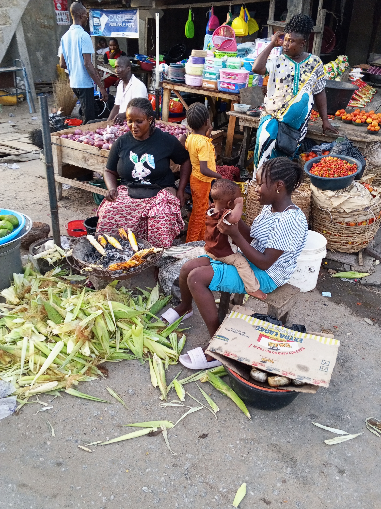 The image depicts an outdoor market scene in Zango-ndaji, Nigeria. A woman wearing a black t-shirt with a rabbit design and a patterned red and white skirt sits near a small charcoal grill with roasting corn. Nearby is a large pile of fresh corn husks. A young woman wearing a striped shirt and blue shorts sits on a stool holding a baby. In the background, a woman in a patterned blouse leans over a table laden with produce, including tomatoes and chili peppers. Beside the produce table sits a wooden table piled high with red onions. Several people stand or sit in the vicinity, including a man in a blue shirt. A faded cardboard box labeled "EXTRA LARGE Bread" sits on top of a bucket containing several objects. The ground is uneven and covered with corn husks. In the background are market stalls with various items for sale, including plastic containers and utensils. A sign reading "CASH AVALABLE HERE" is visible.