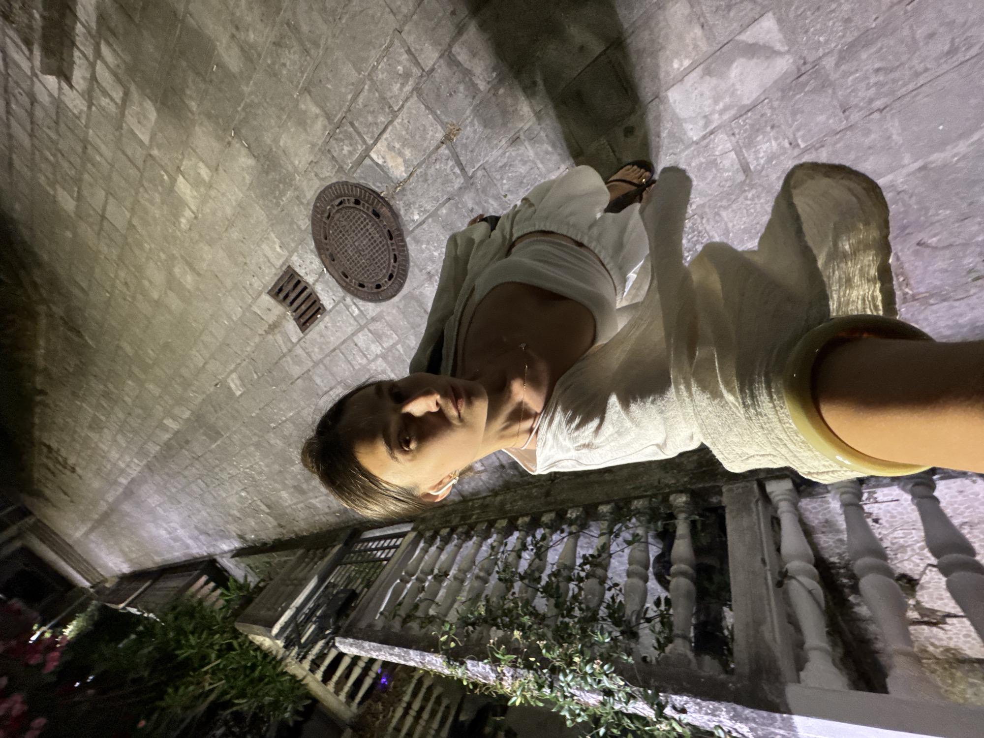 An overhead, full-body selfie is taken by a woman at night in Budva, Montenegro. She stands on a stone-paved walkway next to a stone wall with an ornate balustrade adorned with climbing plants. The woman is wearing a white cropped top, white linen pants, and a light-colored shawl draped over her shoulders. She is wearing sandals on her feet. In the background, there is a large brick wall, a storm drain, and a manhole cover on the ground. The lighting in the image is dim, with artificial lights illuminating the scene.