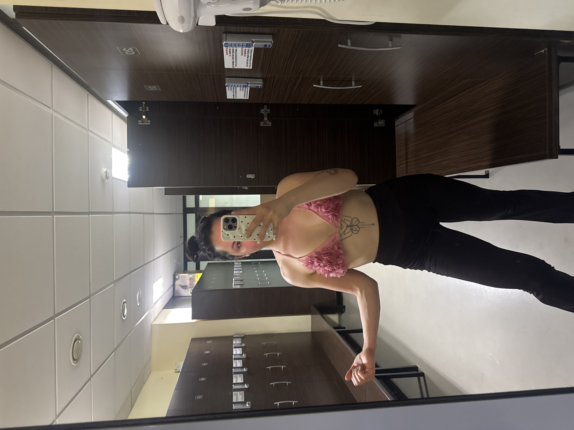 The image captures a young woman taking a selfie in what appears to be a changing room or locker room, possibly in Burgas, Bulgaria. The photograph is taken in a mirror, reflecting the woman and her surroundings.
The subject is a woman with dark hair pulled back, wearing a pink, floral-patterned sports bra or crop top and black leggings. She holds a white smartphone with a speckled case, using it to capture her reflection. A tattoo, resembling a floral or butterfly design, is visible on her left ribcage. Her left arm is bent, and her hand is fisted, suggesting a pose of strength or readiness.
The setting consists of dark wooden lockers with silver-toned handles, lining the walls. Some lockers feature small white cards or displays, though any text on them is too indistinct to read. Faint square symbols are also visible on the locker doors. The walls are a light, possibly tiled, material, and the white ceiling is fitted with multiple recessed circular lights. A white, wall-mounted dispenser or appliance is partially visible in the upper right. The bright lighting indicates an indoor public facility, likely a gym or sports center. The woman's activity is a self-portrait, possibly taken before or after a fitness session.