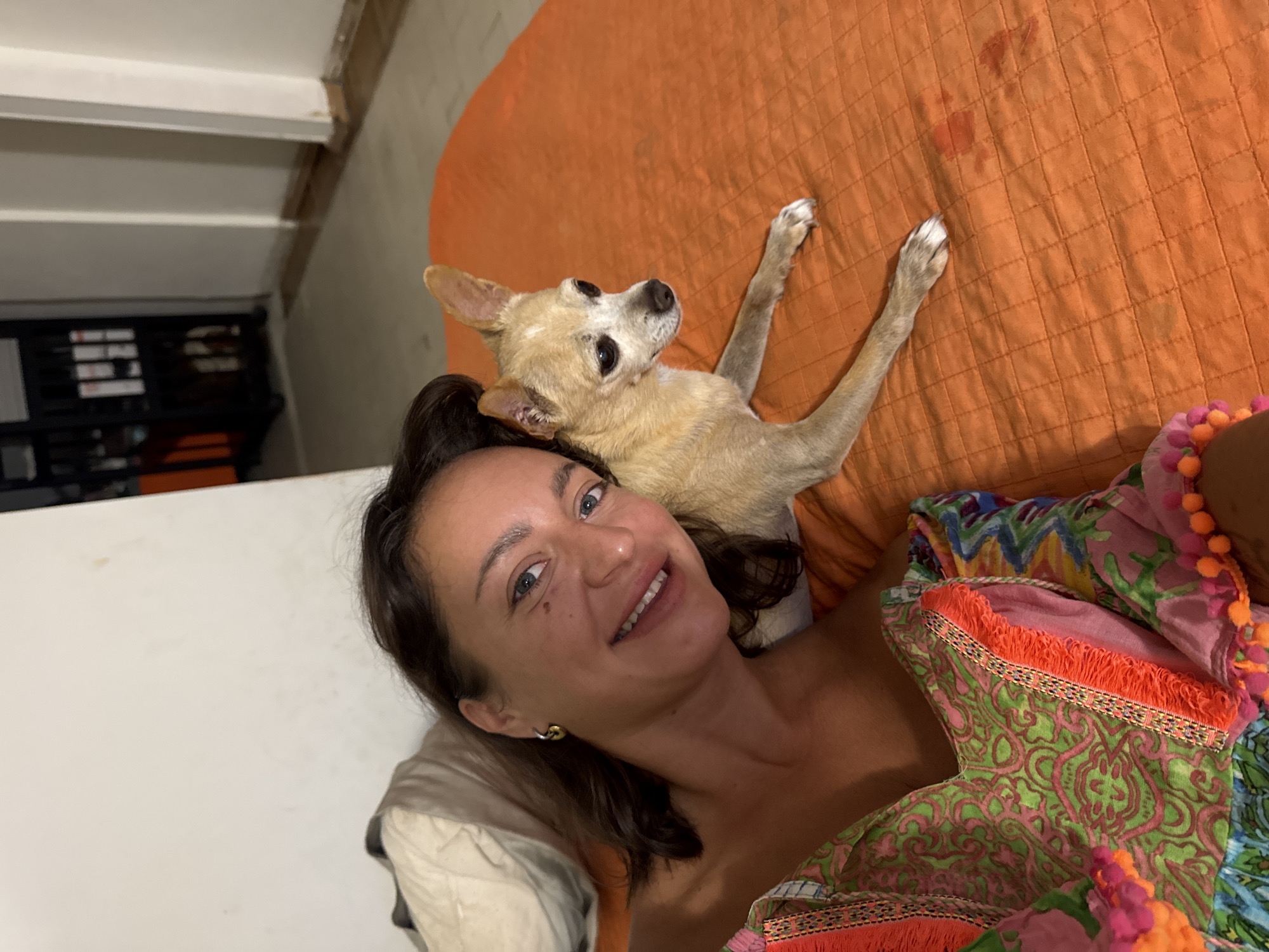 In this close-up, high-angle shot, a woman and her dog are lying on an orange quilted surface, possibly a bed or couch. The woman is in the foreground, smiling at the camera with her head tilted slightly. She has dark brown hair, blue eyes, and is wearing a strapless top with a vibrant, patterned fabric featuring green, pink, and orange accents, including fringed trim and pom-poms. A delicate gold earring is visible on her left ear.
Resting on her chest and head is a small, light-colored Chihuahua with large, dark eyes and erect ears. The dog is looking up and to the right, its front paws extended. The background shows a white surface on the left, possibly a wall or furniture, and a darker area with shelving and some items on the right, hinting at an indoor setting. The overall lighting suggests it could be daytime or a well-lit indoor space. The mood appears relaxed and affectionate, given the close proximity and smiles of both subjects. The setting is likely domestic. No visible text is present in the image.