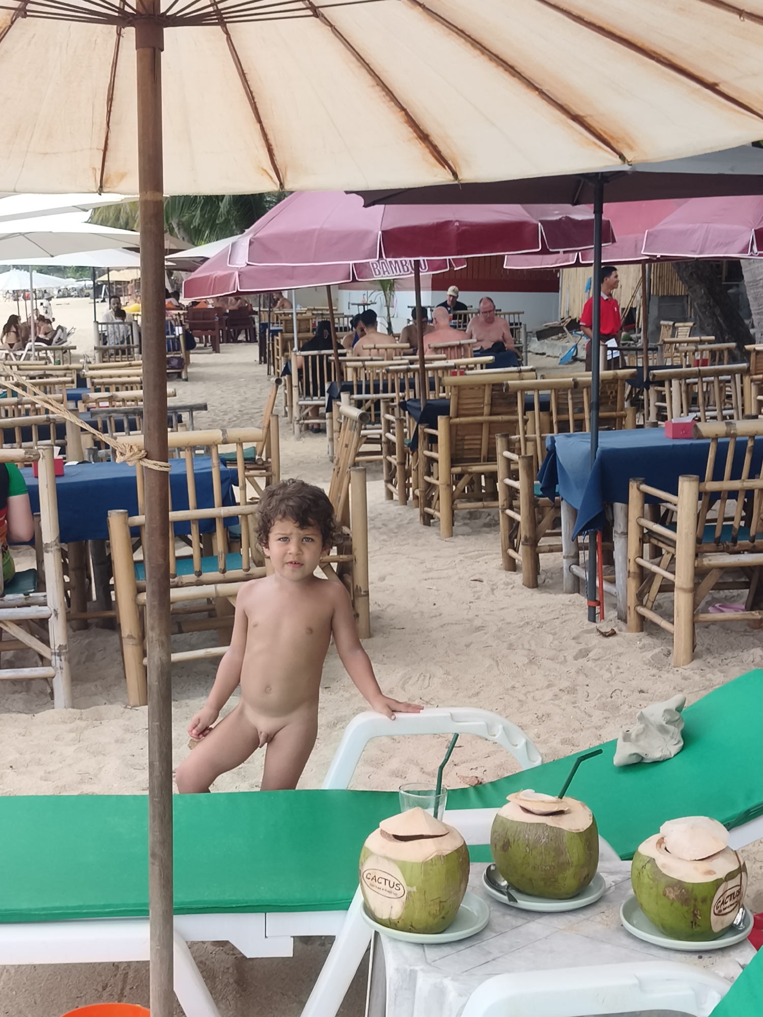 This media file captures a scene on a sandy beach in Baan Lamai, Thailand. The foreground features a young, unclothed boy with curly hair, standing and looking towards the camera. He is positioned near a green lounge chair and a table adorned with three coconuts, each with a straw and a small lid. One of the coconuts has the word "CACTUS" visible on it.
In the background, a beachside restaurant is set up with numerous tables and chairs made of bamboo. Several people are seated at these tables, some appearing to be patrons and others possibly staff. Two large umbrellas, one pale and weathered and another a darker burgundy, provide shade over the seating area. The overall atmosphere suggests a casual, daytime setting, likely warm and sunny given the beach location and the presence of umbrellas. The sandy ground, the palm trees visible in the distance, and the open-air dining setup further reinforce the tropical beach environment. There is no indication of specific emotions displayed by the people, beyond a general sense of relaxation associated with a beach vacation.