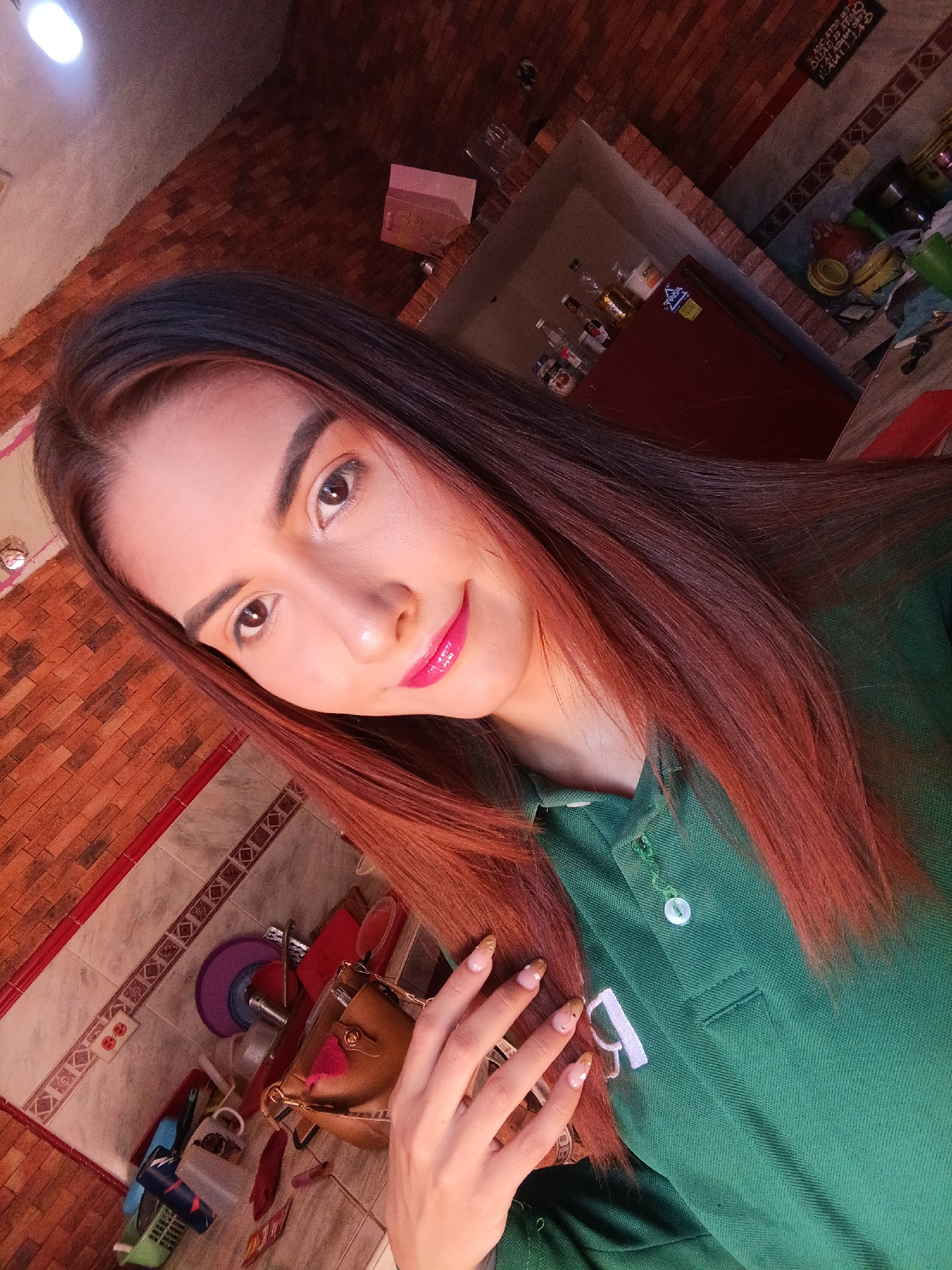 A young woman with long, reddish-brown hair and bright pink lipstick is posing for a selfie. She is wearing a green polo shirt. Her left hand is raised, displaying manicured nails with a light base and gold accents.
The background is a cluttered domestic setting. A section of brick wall is visible, along with tiled surfaces. Behind the woman and to the right, is a counter with bottles and miscellaneous items on a shelf or in a recess. Below and to the left of the woman is a cluttered countertop holding an open purse, and various colored utensils and containers. It is not possible to verify the precise location in Juan Griego, Venezuela without further corroborating evidence.