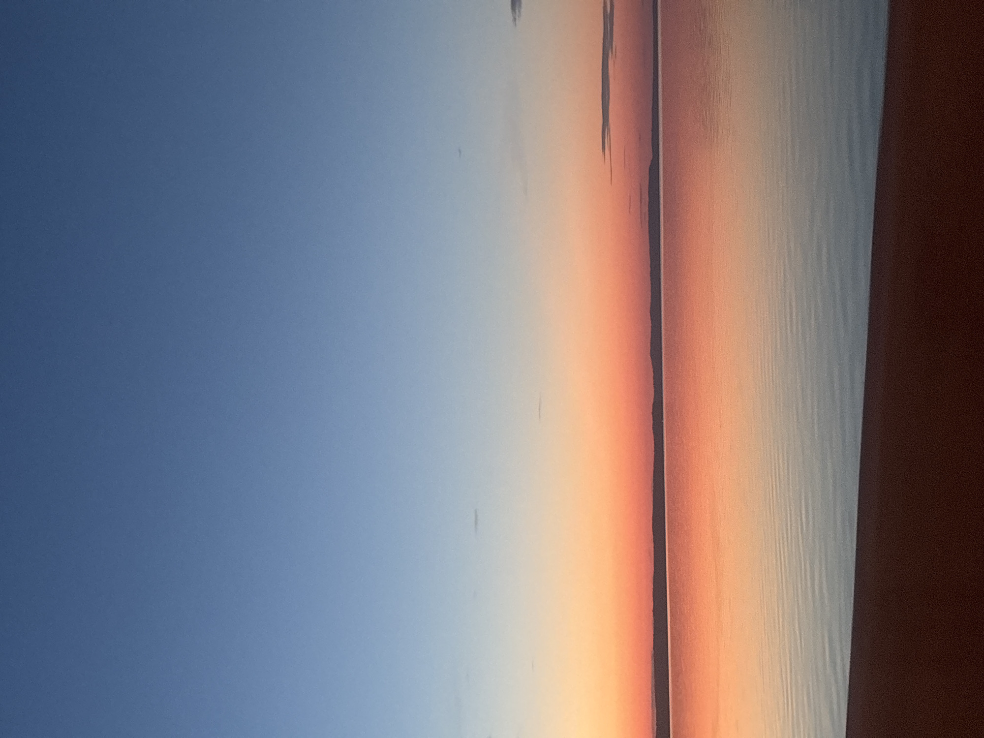 This image, taken in Chaikovsky, Russia, depicts a vast, serene waterscape at either sunset or twilight. The upper portion of the sky is a clear, deep blue, gradually transitioning downwards to a wide band of vibrant orange and red hues near the horizon. A thin, dark, undulating line, indicative of distant land or hills, separates the colorful sky from the water. The expansive body of water perfectly mirrors the sky's gradient, displaying warm orange and red reflections closer to the horizon, which then transition into cooler, lighter blue and grey tones in the mid-ground. Subtle, horizontal ripples are visible across the water's surface, suggesting calm conditions. The immediate foreground features a dark, reddish-brown strip of land, likely a beach or bank, occupying the bottom segment of the frame. No people, animals, or artificial structures are discernible within the scene.
