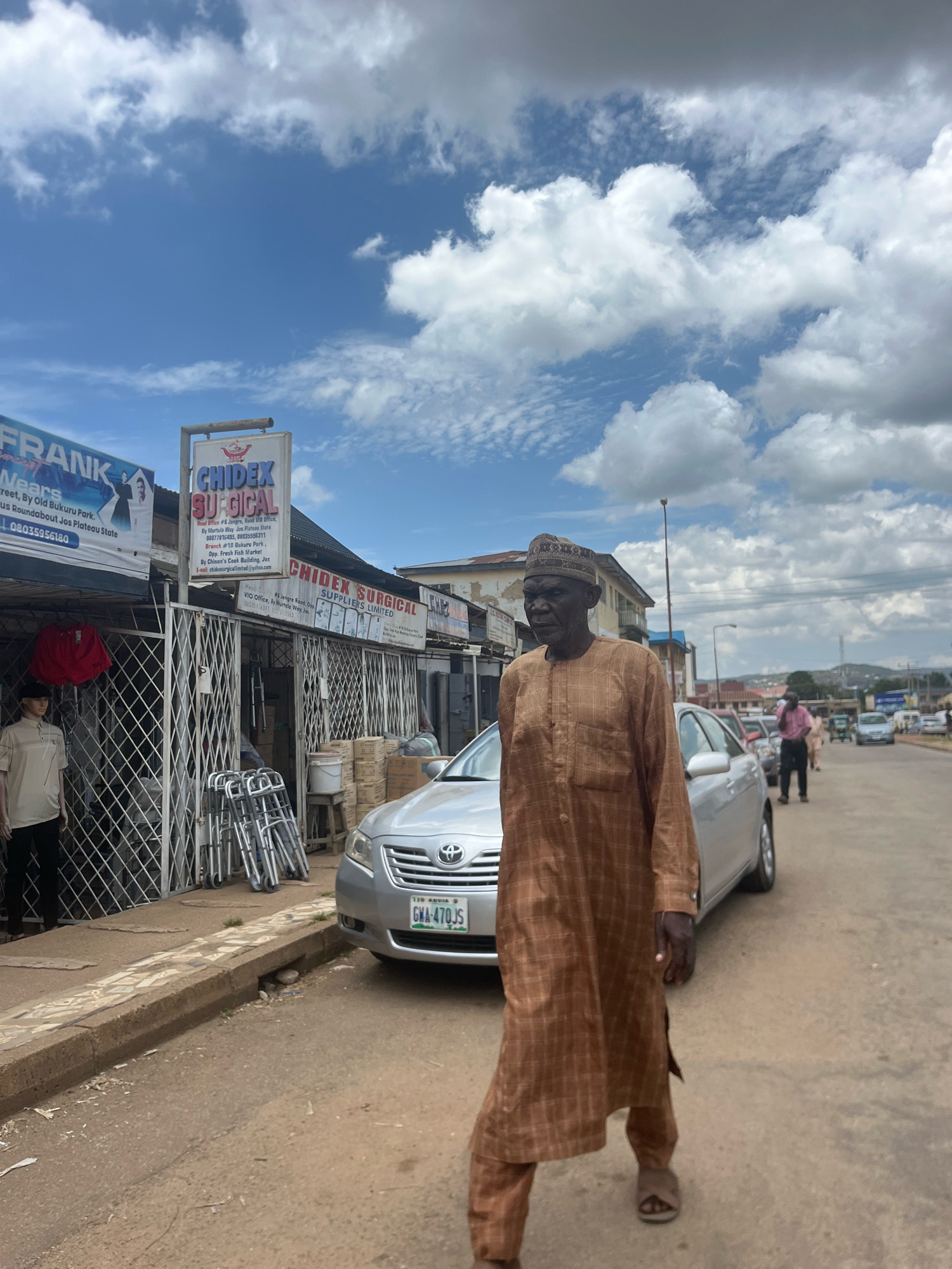 The image captures a street scene in Jos, Nigeria, with a man in the foreground walking towards the viewer. He is wearing a brown, checkered traditional outfit and a patterned cap. Behind him, a silver sedan with a Nigerian license plate is parked on the side of the road. To the left, several storefronts are visible, including one with a sign for "CHIDEX SURGICAL." These shops appear to be on a commercial street. In the background, other people are walking along the street, and vehicles are moving in the distance. The sky is partly cloudy with dramatic white cumulus clouds against a blue background. The overall atmosphere suggests a typical day in a bustling Nigerian city.