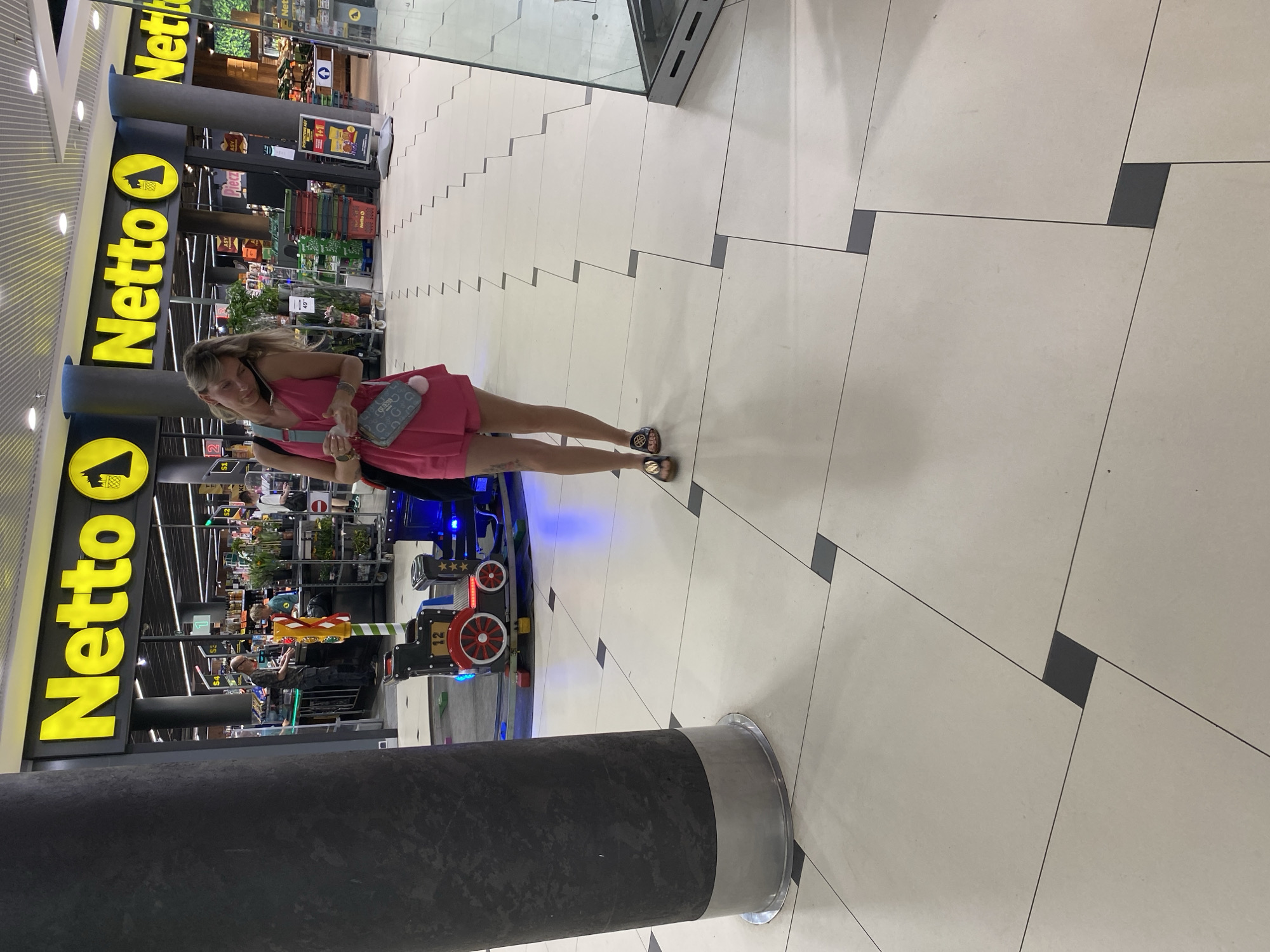 A woman in a bright pink dress stands in front of a "Netto" grocery store. She is looking down and holding a light blue purse with a grey pom-pom. Her right foot is raised slightly, and she wears dark sandals. The background shows the interior of a well-lit store with shelves stocked with products and a children's toy train ride. The floor is tiled with a pattern of light and dark squares. Visible text includes "Netto" in large yellow letters on a black background, and other signage within the store. The scene appears to be indoors, likely in a shopping mall or the vestibule of a supermarket. The lighting suggests daytime.