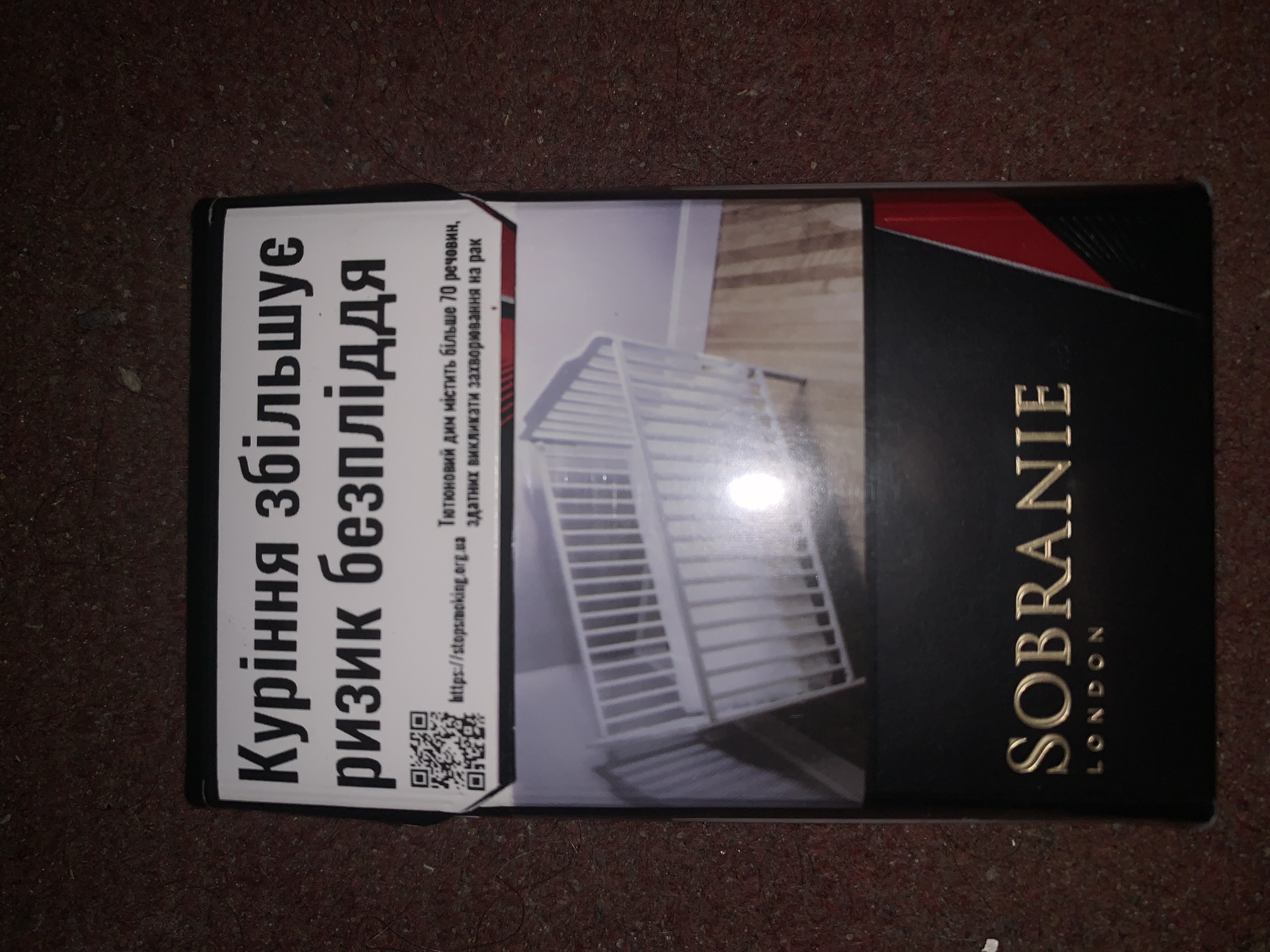 The image shows the front of a pack of Sobranie cigarettes. The pack is black with gold lettering and has a graphic warning about the dangers of smoking. The warning text is in Ukrainian and reads "Smoking increases the risk of illness." Below the warning text is an image of an empty crib. The bottom of the pack features the brand name "SOBRANIE" in gold lettering, with "LONDON" below it. The background of the image is a dark, textured surface.