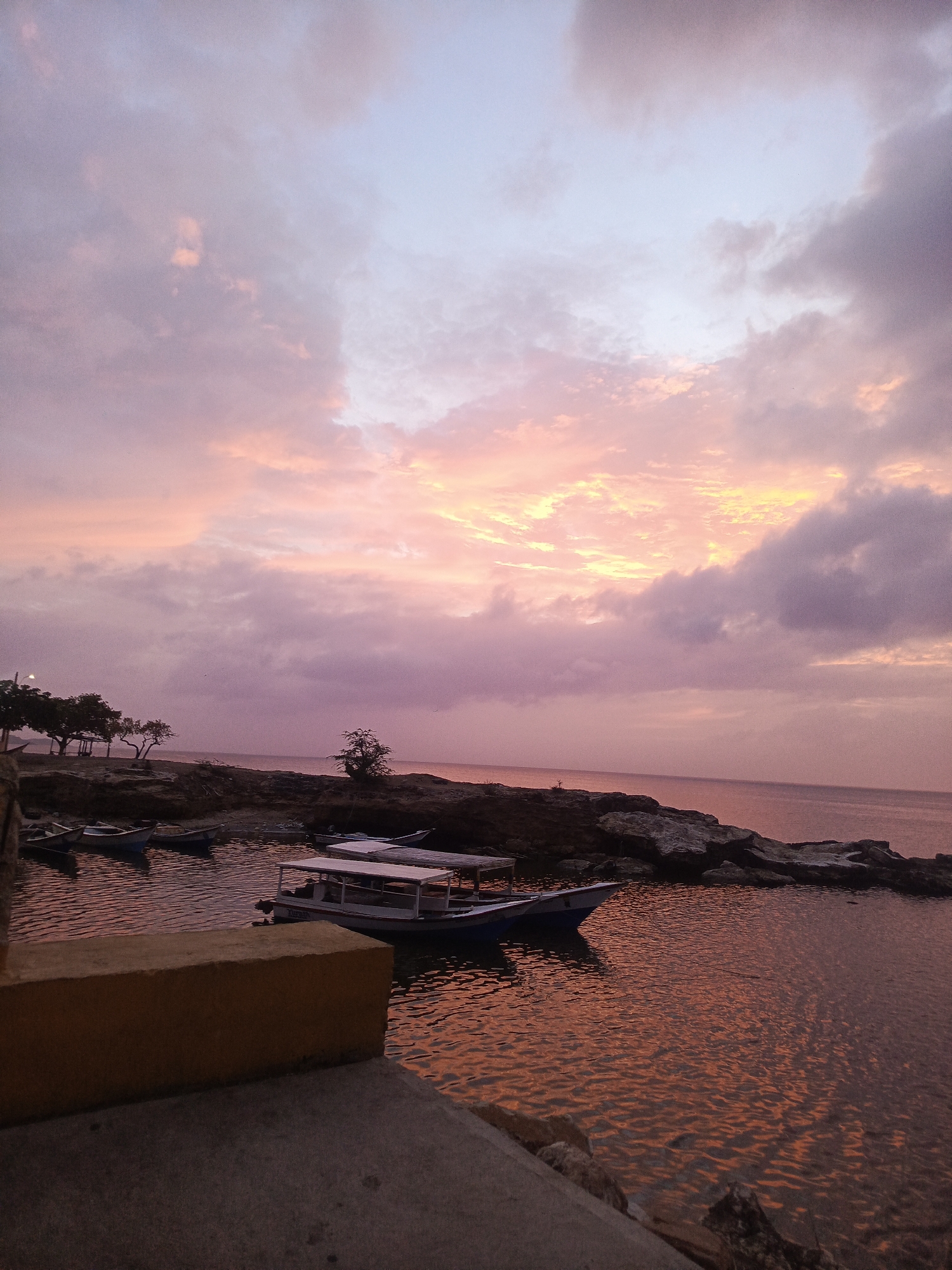 The image depicts a sunset scene in Juan Griego, Venezuela. The sky is filled with colorful clouds, transitioning from pink to purple. Below the sky, the sea reflects the colors of the sky, with visible water ripples. Several small boats are docked in a calm bay, with rocky landmasses in the distance. Trees and buildings line the shoreline. A yellow concrete wall is in the foreground, adding depth to the image. The overall scene suggests a peaceful and serene coastal environment.