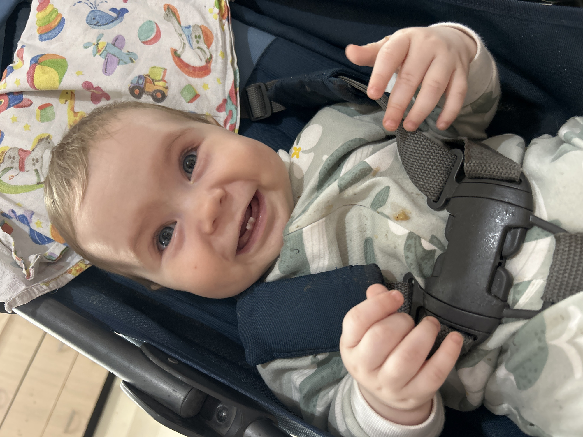 A baby lies in a stroller or car seat, secured with a grey harness. The baby is wearing a patterned onesie with light green and white foliage. The baby is smiling and has two visible teeth. The baby has blonde hair and blue eyes. A patterned blanket with cartoon-style toys is visible to the left of the baby's head. The stroller or car seat is dark blue. The location is unknown and there is no mention of Rusalevka, Russia.