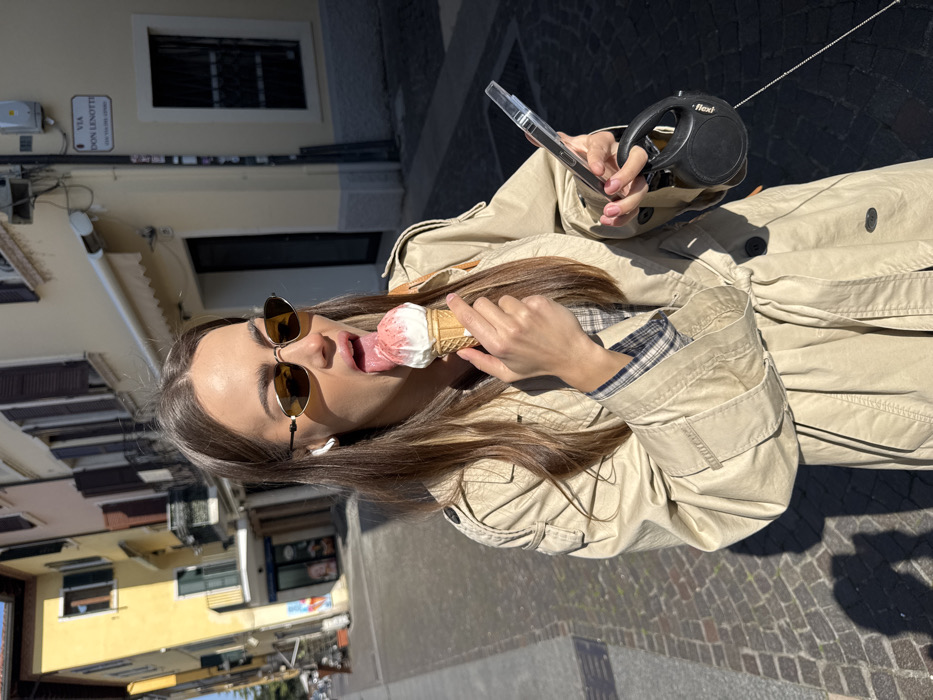 A young adult female is depicted on a cobblestone street, identified as Via Don Lenotti in Peschiera del Garda, Italy, under bright sunlight. She has long brown hair and is wearing oval-shaped gold-rimmed sunglasses and a beige trench coat over a plaid shirt. A white earbud is visible in her left ear. Her mouth is open with her tongue extended, actively licking a two-scoop ice cream cone held in her right hand; the top scoop is pink with red specks, and the bottom is white. In her left hand, she holds a mobile phone encased in clear plastic and a black Flexi brand retractable dog leash. Distinct shadows are cast on the cobblestone pavement from the sunlight originating from the left. Buildings with various light-colored facades and dark-shuttered windows line the street in the background.