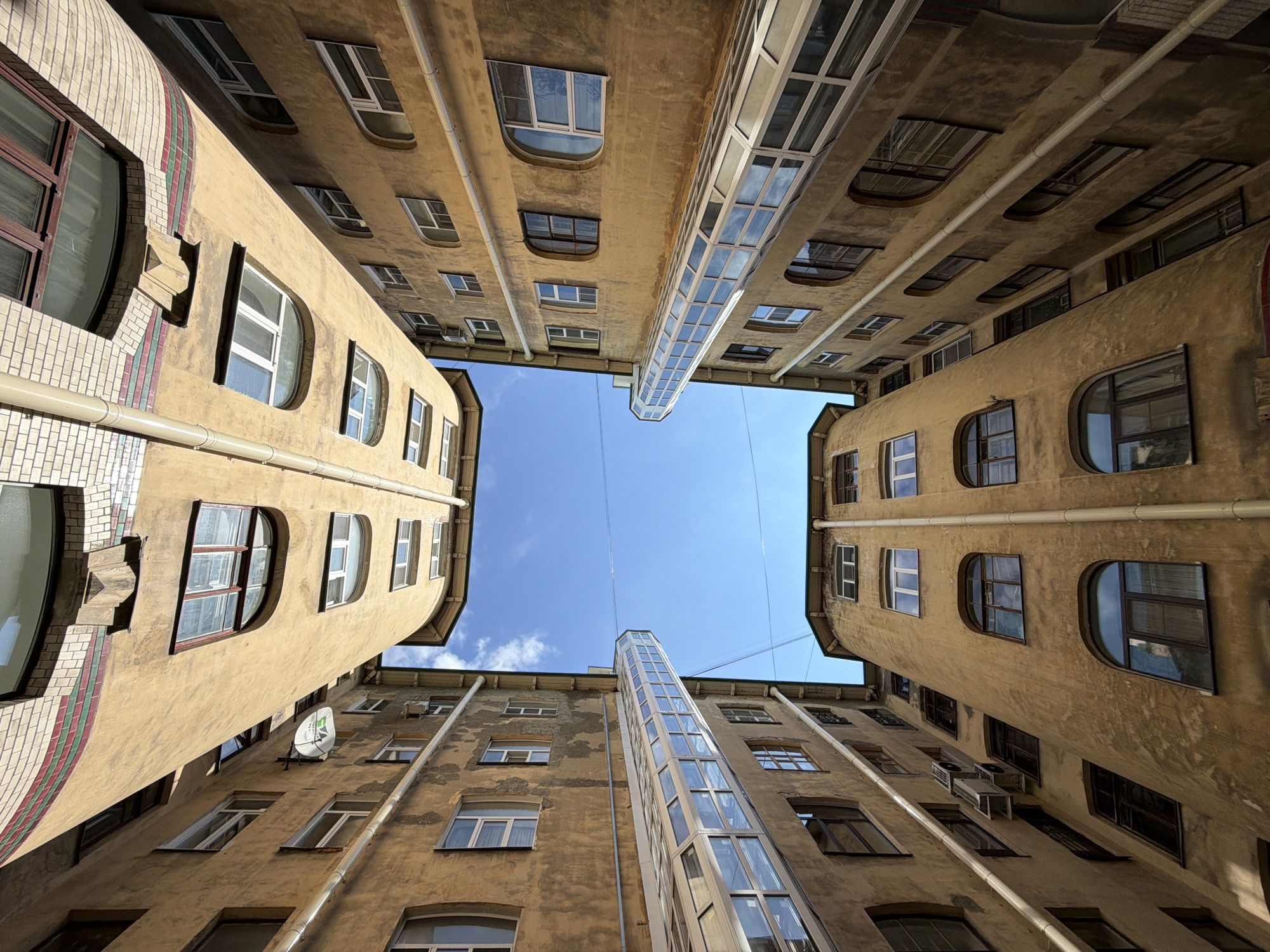 The image captures an extreme upward-angle perspective from within a narrow, enclosed courtyard surrounded by multi-story buildings in Saint Petersburg, Russia. The perspective creates a converging effect, emphasizing the height of the structures and the limited expanse of the sky.
Four distinct building facades are visible, characterized by their light brown or tan, textured stucco finish. These buildings show signs of age, including weathering and discoloration. Various windows, including rectangular, arched, and rounded-top designs, are scattered across the facades, some with dark frames and others with white.
A prominent feature includes two enclosed, glass-paneled sky bridges or walkways spanning the courtyard high above. These modern structures connect the buildings, with their glass surfaces reflecting the bright, clear blue sky, which contains a few wispy white clouds.
Other architectural details include vertical drainage pipes along the building exteriors. On the left, a curved building corner is adorned with decorative brickwork in red, green, and white stripes, featuring a small, ornate corbel. A small white satellite dish is mounted on one wall, and an external air conditioning unit is visible on another.
The photograph depicts a bright, clear daylight scene, likely midday or early afternoon. No people, animals, or vehicles are present, placing sole focus on the urban architecture. The overall impression highlights the dense, historical building styles characteristic of Saint Petersburg's unique courtyards. No discernible text is clearly visible, though a logo appears on the satellite dish.