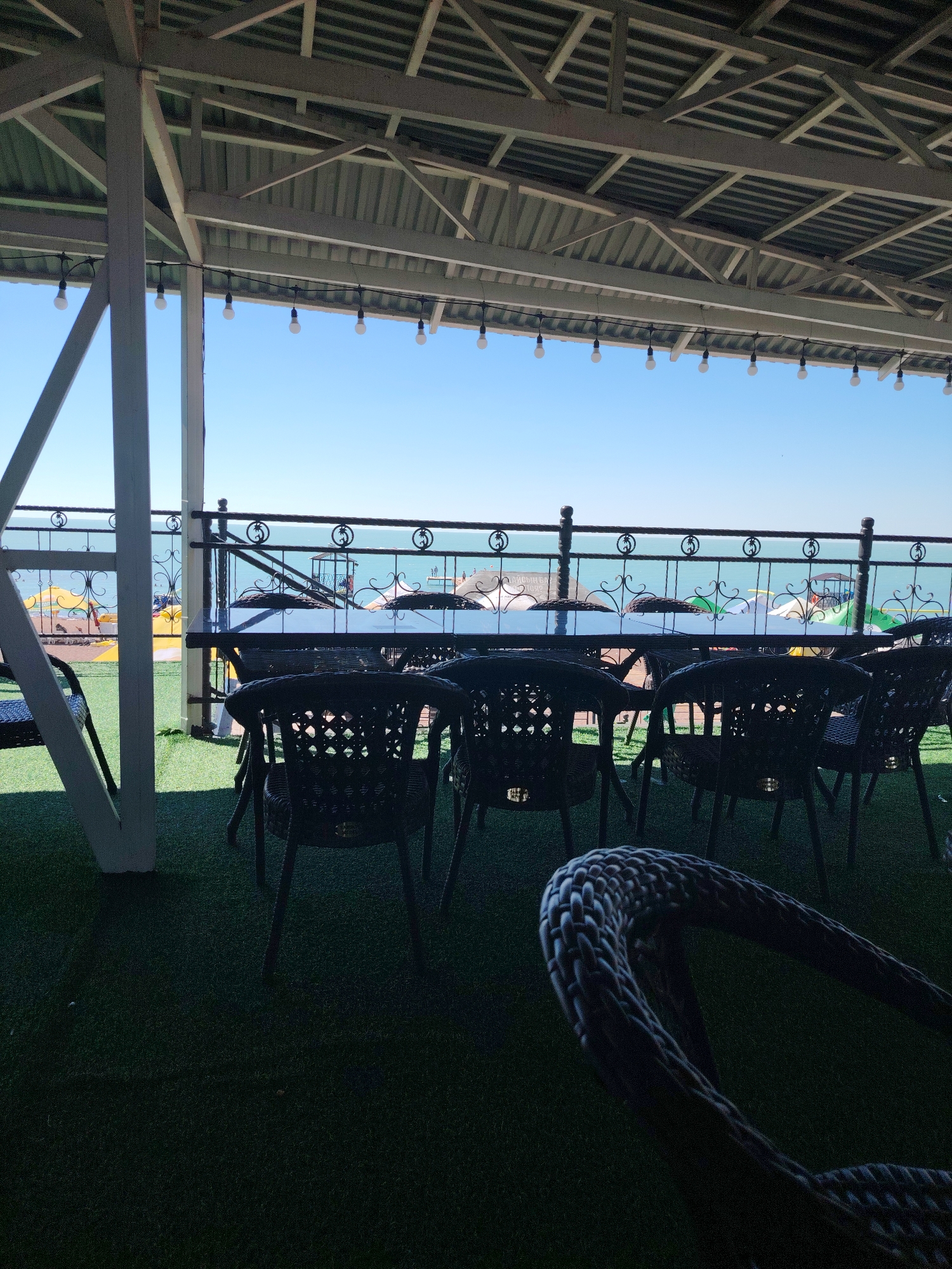 The image shows an outdoor seating area at a cafe or restaurant on the shore of Lake Balkhash in Toranghylyq, Kazakhstan. The foreground features dark wicker chairs and a table, with a braided rope chair element in the lower right corner. The seating area is situated on a green artificial turf surface, possibly Astroturf. Beyond the seating, a black metal railing with decorative scrollwork separates the area from the lake. The lake's water is a vibrant turquoise, and the sky is a clear, pale blue. In the distance, across the lake, beachgoers can be seen with yellow umbrellas and inflatable watercraft, indicating a recreational area. The seating area is covered by a light-colored, slatted roof structure, supported by white pillars. A string of small, white globe lights is suspended from the roof. The overall impression is of a relaxed, seaside dining or lounging establishment.