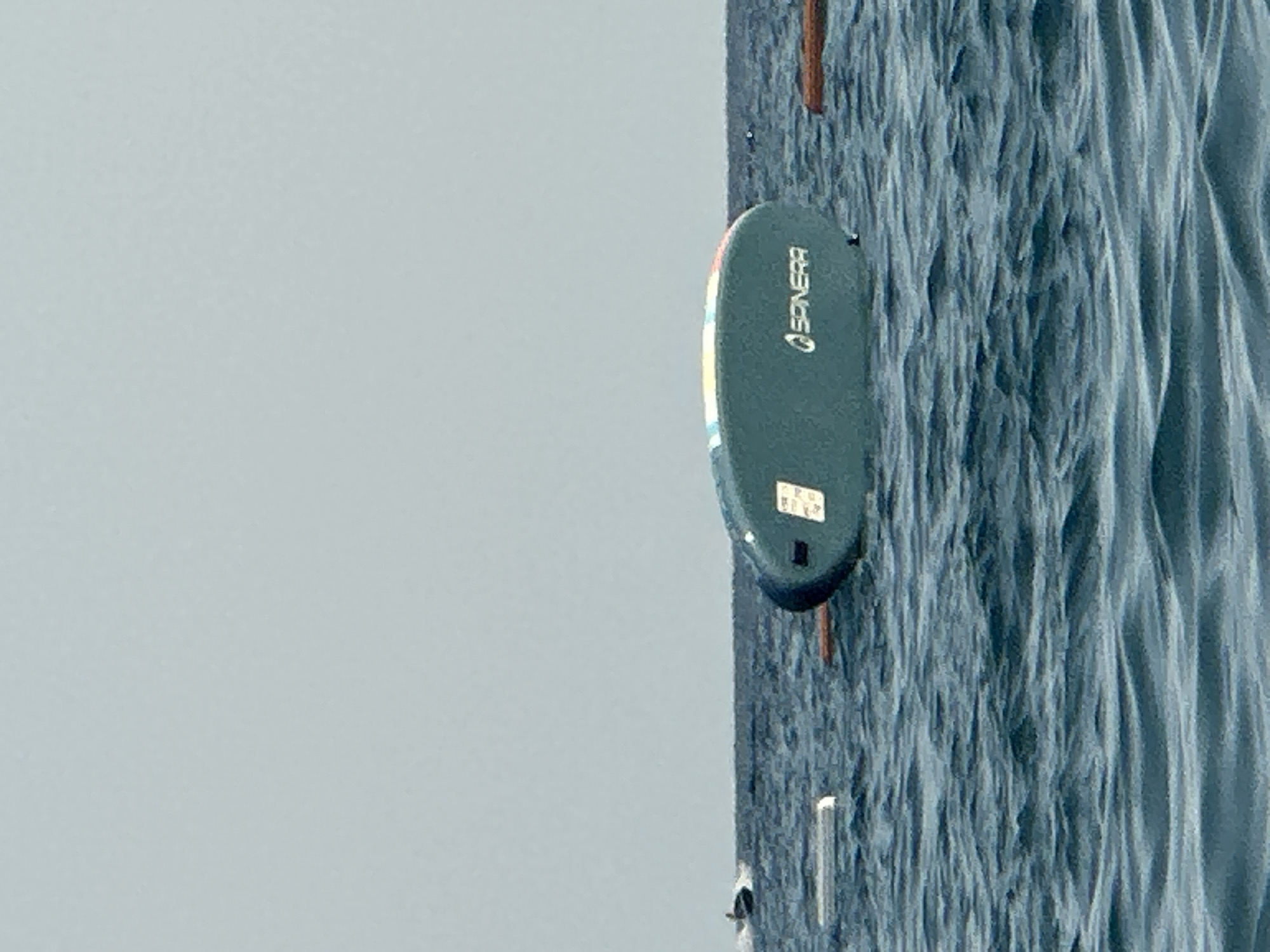 A teal-colored paddleboard with colorful accents and the logo "SPNERRA" is floating on its side in the water. A white rectangular object is next to it, also floating. In the distance, a person is riding a jet ski. The scene takes place on the water with a clear, light blue sky in the background. The location is the Bay of Kotor, near Boreti, Montenegro.