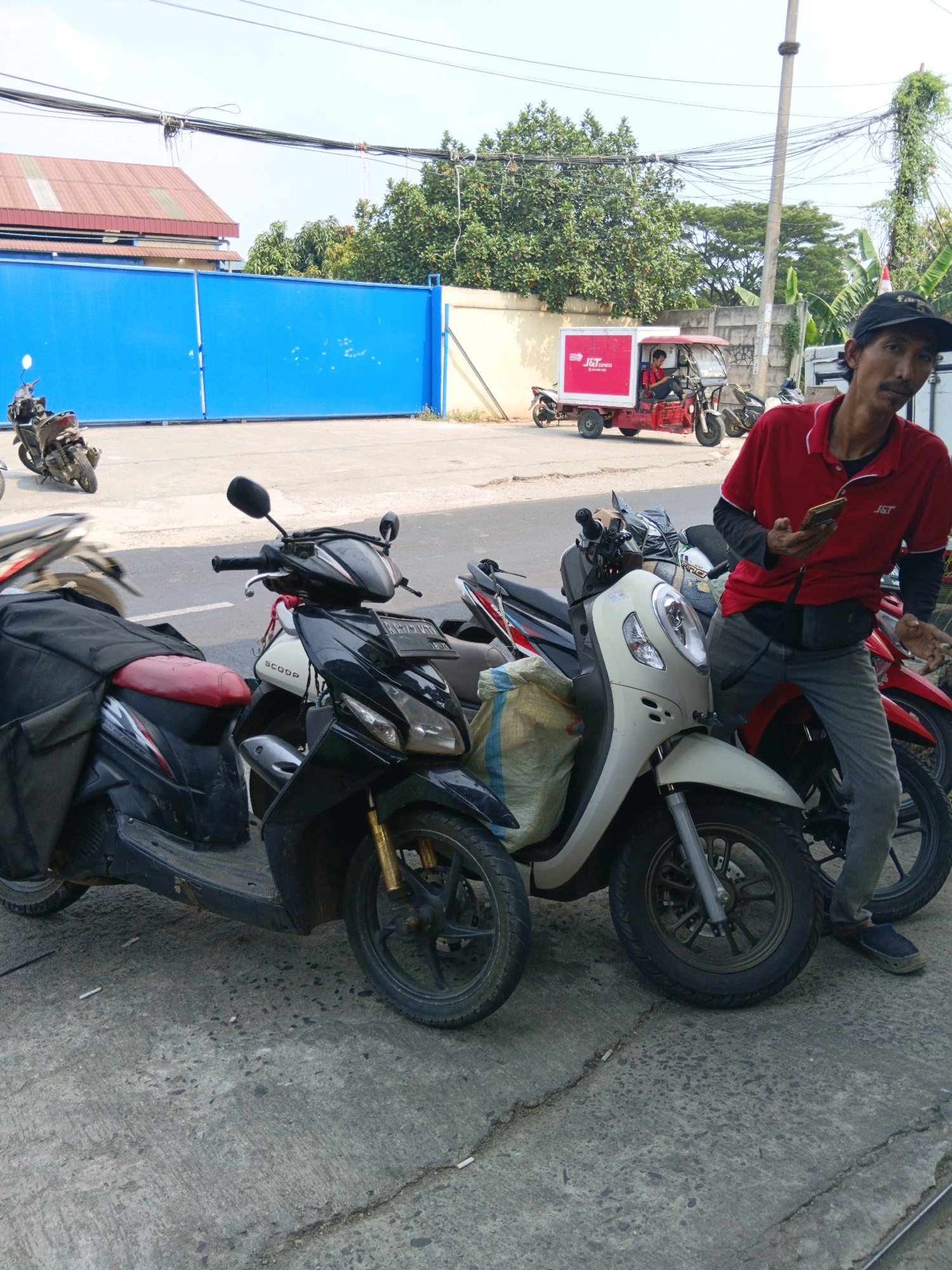 In Bekasi, Indonesia, a man in a red polo shirt with a J&T Express logo stands near several motorcycles. He holds a smartphone in his right hand and appears to be looking at it. The man is positioned in front of a cream-colored scooter and to the left of a red scooter. Behind him, a red J&T Express delivery vehicle is parked. The environment includes a large blue gate, buildings with red roofs, trees, and utility poles with numerous wires. The ground is paved with concrete.