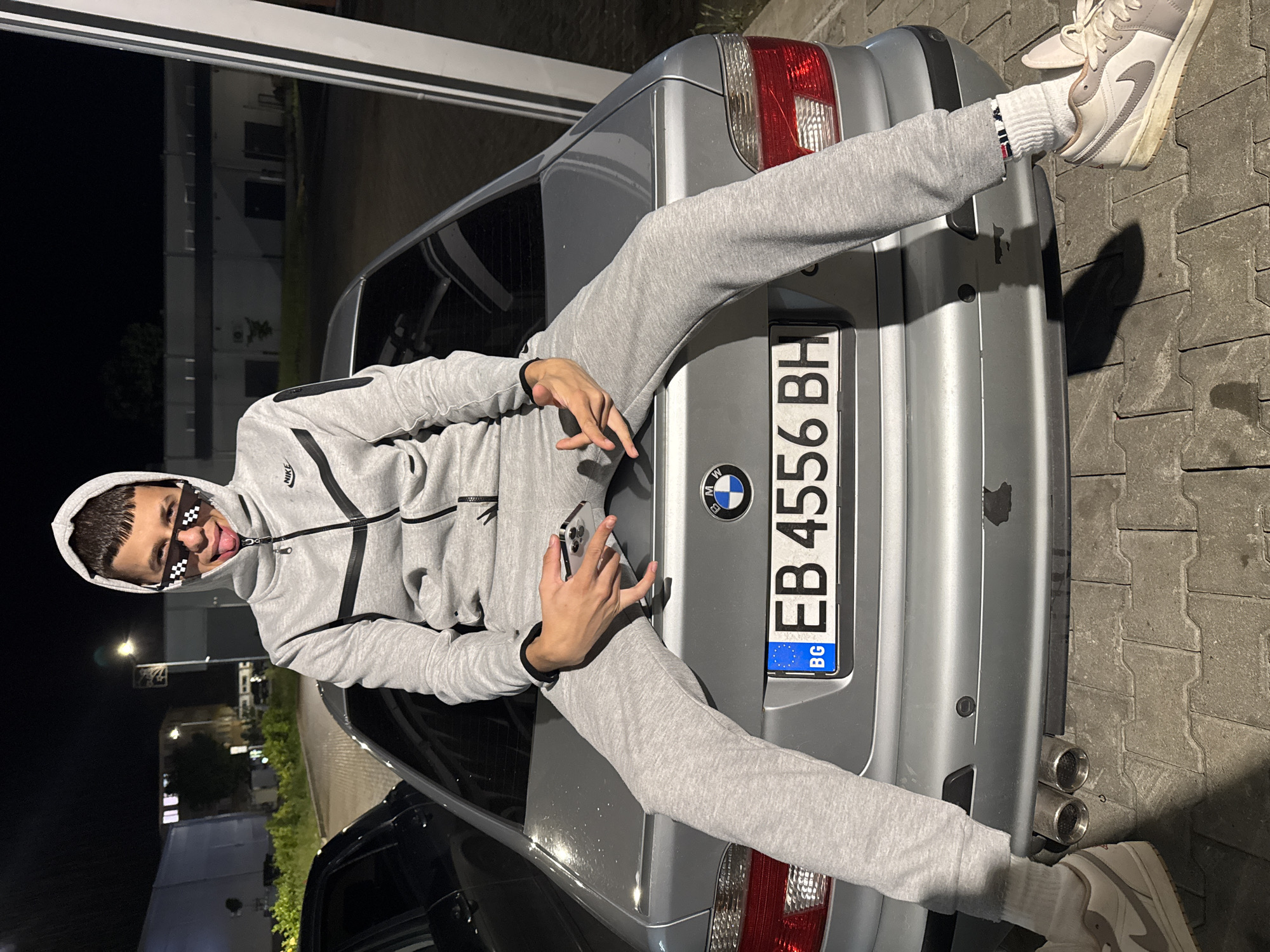 A young man poses playfully on the trunk of a silver BMW sedan at night in what appears to be a parking lot or paved area in Sevlievo, Bulgaria. He is positioned with his legs spread over the car's trunk, looking directly at the camera with a smile and his tongue sticking out. He wears a grey Nike Tech Fleece tracksuit, with the hoodie pulled up, and pixelated "deal with it" sunglasses. His left hand is raised in a gesture, possibly a peace sign, while his right hand holds a smartphone. He wears white socks with subtle red and blue stripes and light-colored Nike sneakers, likely Air Jordan 1s.
The car is a silver BMW, possibly a 5-series, with a visible BMW emblem on the trunk. Its Bulgarian license plate reads "EB 4556 BH," where "EB" indicates the Gabrovo region, which aligns with the provided location of Sevlievo. Dual exhaust pipes are visible on the right side of the car's rear bumper. The background is dark, indicating nighttime, with distant artificial lights and blurry outlines of buildings. The ground beneath the car is paved with block-like patterns. The overall mood is casual and youthful.