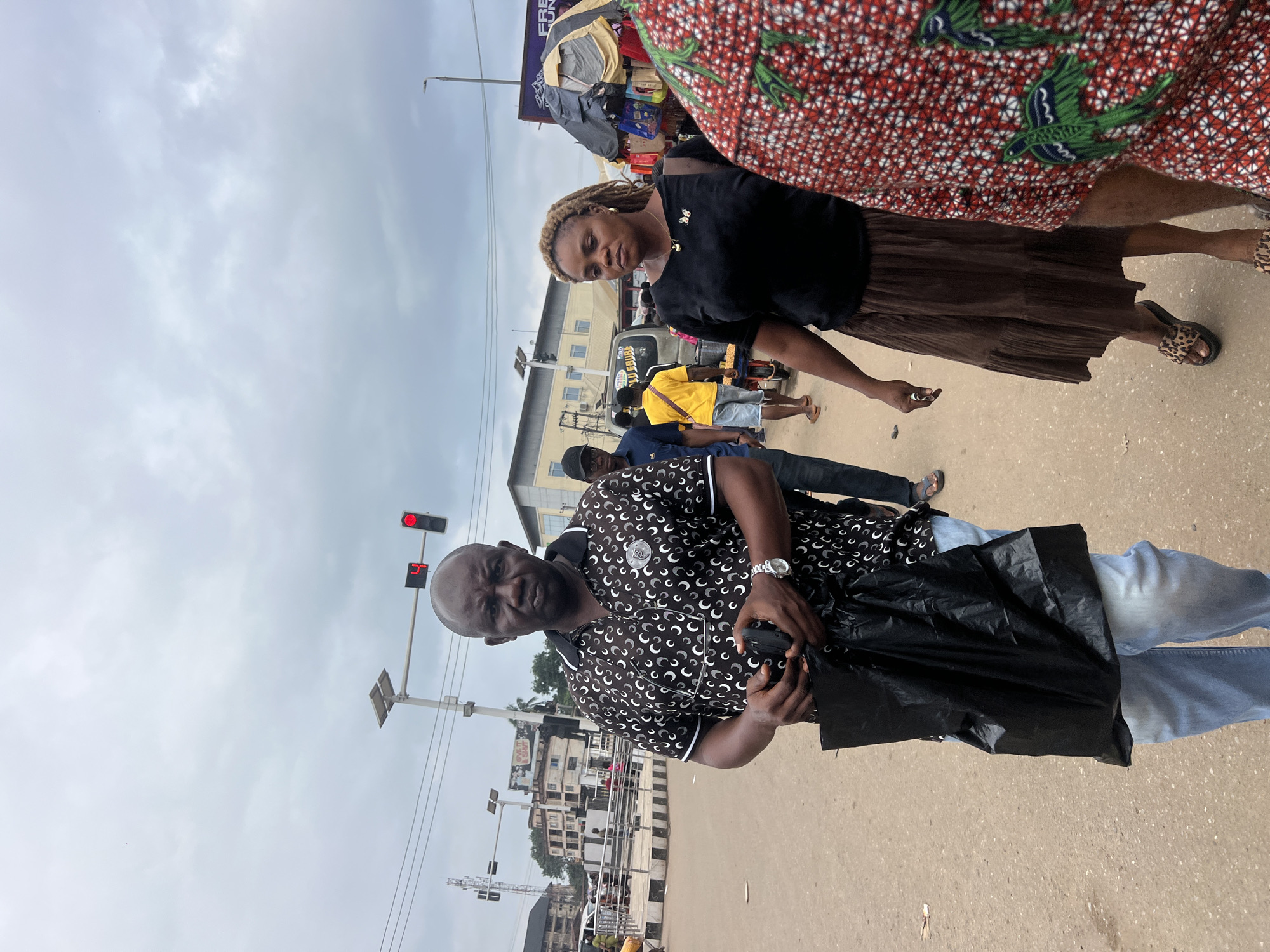 A medium shot captures a street scene in Nkpor, Nigeria, under an overcast sky. In the foreground, a man with dark skin and a shaved head, wearing a black short-sleeved polo shirt with a white circular pattern, holds a black plastic bag in his left hand and a dark object, possibly a phone, in his right. He is looking directly at the camera with a serious expression. To his right, a woman with braided blonde hair, wearing a black t-shirt and a brown tiered skirt, walks past. Her gaze is directed slightly away from the camera. Behind them, other people are visible, including a person in a yellow shirt and denim shorts in the middle ground, and a person in a red and green patterned garment in the right foreground. Traffic lights with a red signal are visible in the background, along with buildings and power lines. The ground appears to be a paved road.