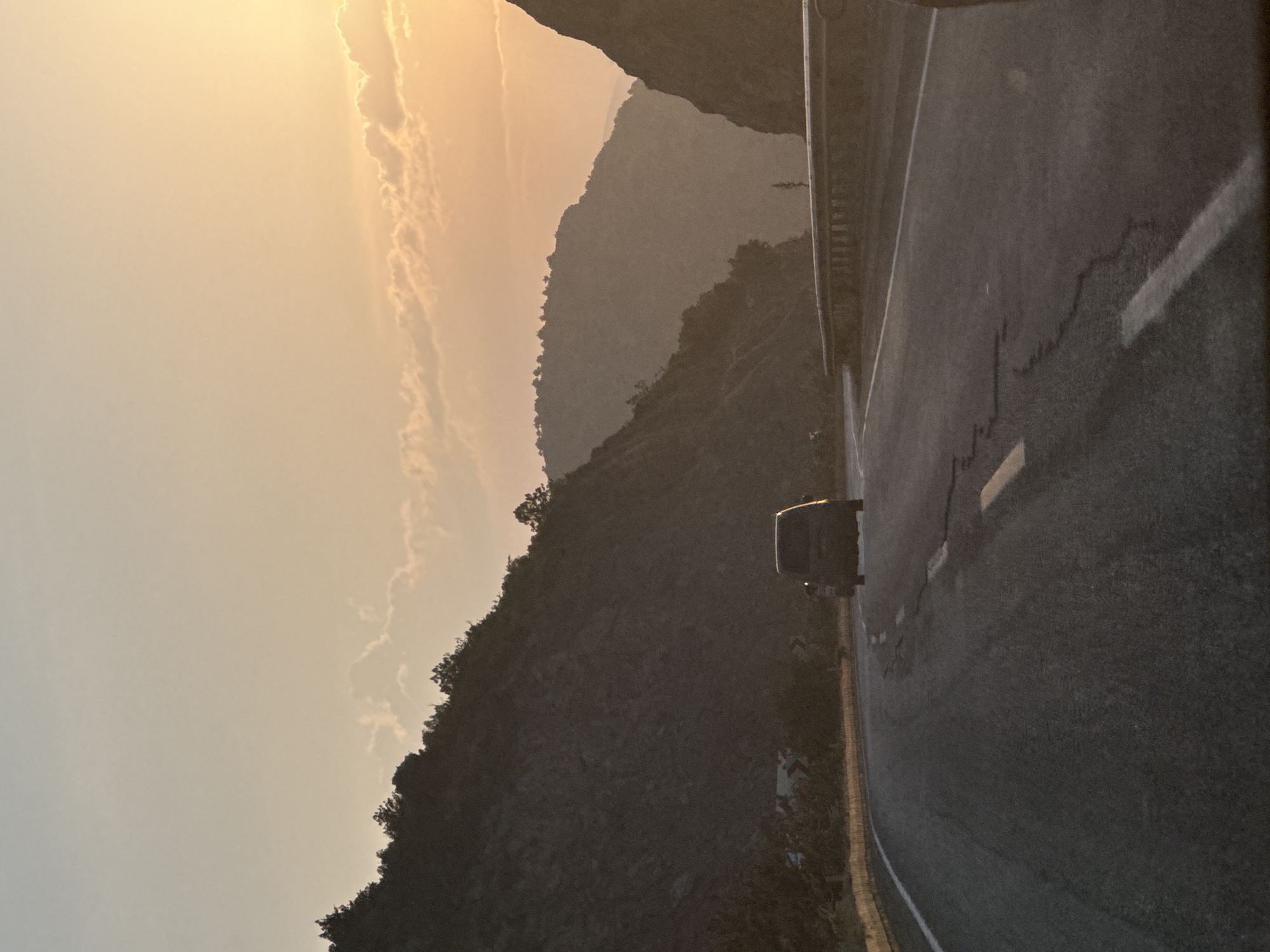The image depicts a mountain road near Korkuteli, Turkey, during either sunrise or sunset. The scene is dominated by a clear asphalt road curving to the right through a mountainous landscape.
The sky in the upper left transitions from a pale blue to a warm, bright orange and yellow hue in the upper right, indicative of a low sun just outside the frame. Scattered, elongated clouds are visible, particularly in the warmer areas of the sky.
The road features white lane markings, some of which exhibit cracks and wear. A thin yellow barrier or curb lines the left edge of the road in the foreground. Further along the right side of the road, a grey metal guardrail is visible. A dark-colored vehicle, resembling a minivan or SUV, is seen traveling away from the viewer in the mid-ground, silhouetted against the bright sky.
Steep, dark mountain slopes covered in trees flank both sides of the road. The mountain on the left is closer and features a dense tree line along its ridge. On the right, multiple layers of mountains recede into the distance, appearing hazy and dark against the illuminated sky. Two white chevron signs, indicating a right turn, are partially visible on the left side of the road, partially obscured by vegetation. No individuals are discernible in the image.