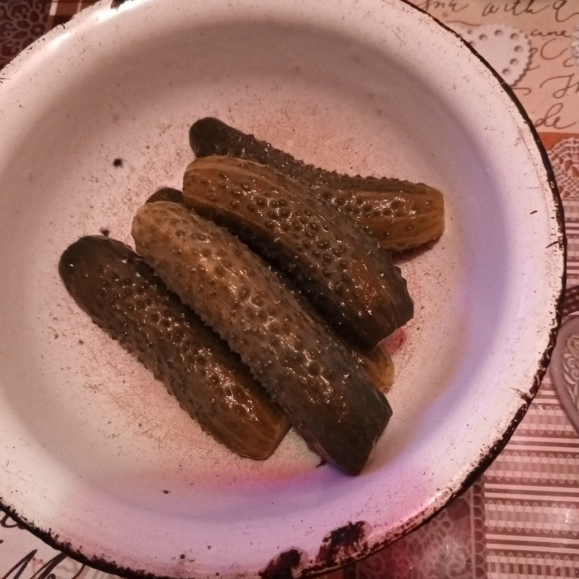 A close-up overhead shot shows a white enamel bowl filled with pickles. The bowl is worn and has dark brown rimming. There are at least five pickles visible, all whole and appearing to be gherkins or cornichons, with bumpy skins. The pickles are a mix of dark green and brownish-green hues, some glistening. They are arranged diagonally across the bowl, with one partially obscuring another. The background is out of focus but appears to be a patterned surface, possibly a tablecloth.