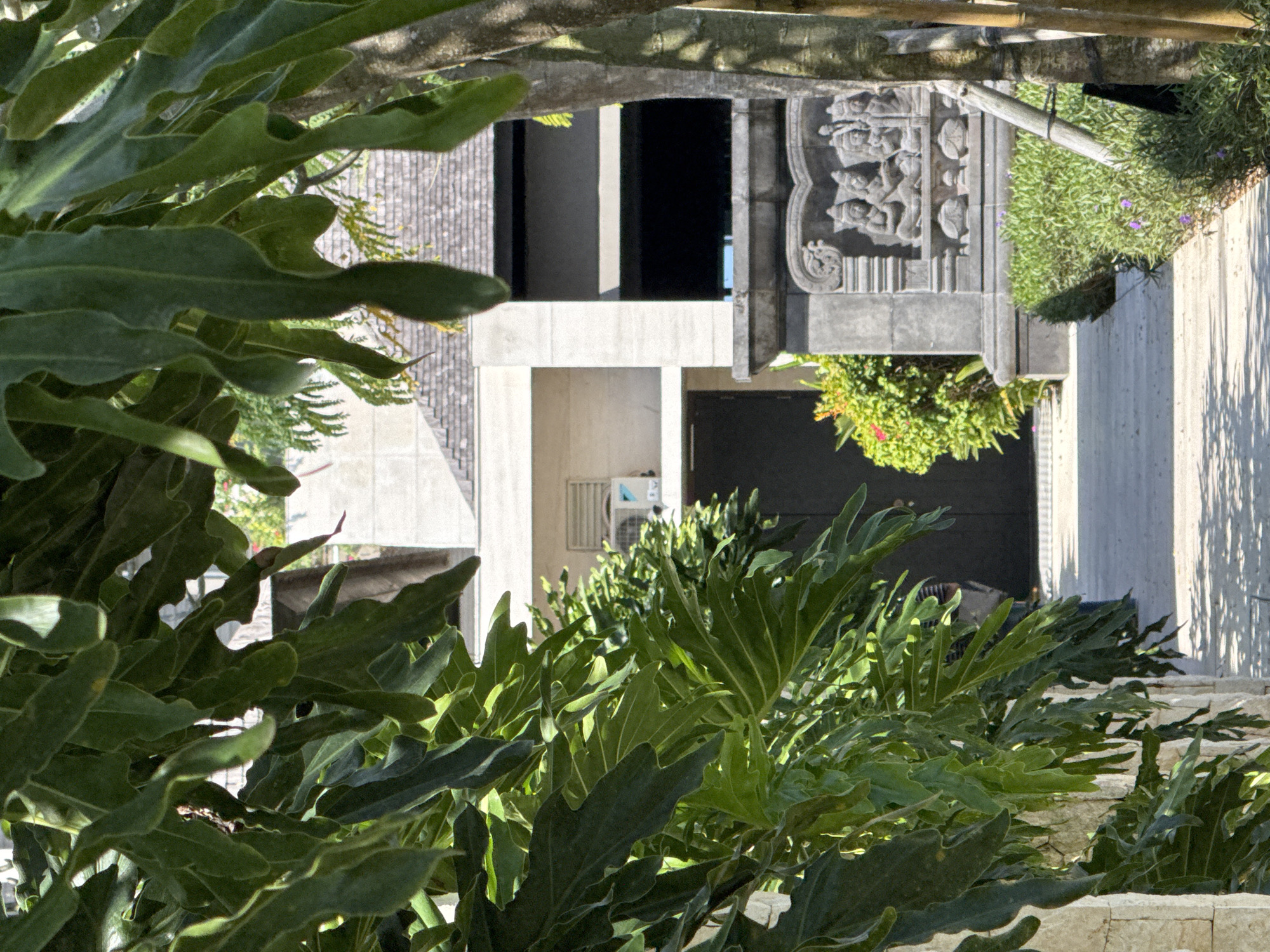 A low-angle, obscured view captures a Balinese architectural scene, likely within the Kuta Selatan area of Indonesia. Lush, green vegetation dominates the foreground, partially concealing the details of a building complex. The building appears to be modern, with geometric shapes and concrete structures. Some architectural details include traditional carvings, possibly depicting deities or mythical figures. The presence of decorative plants and foliage suggests a well-maintained or landscaped environment. The scene appears to be tranquil, devoid of human activity, emphasizing the architectural and natural beauty.