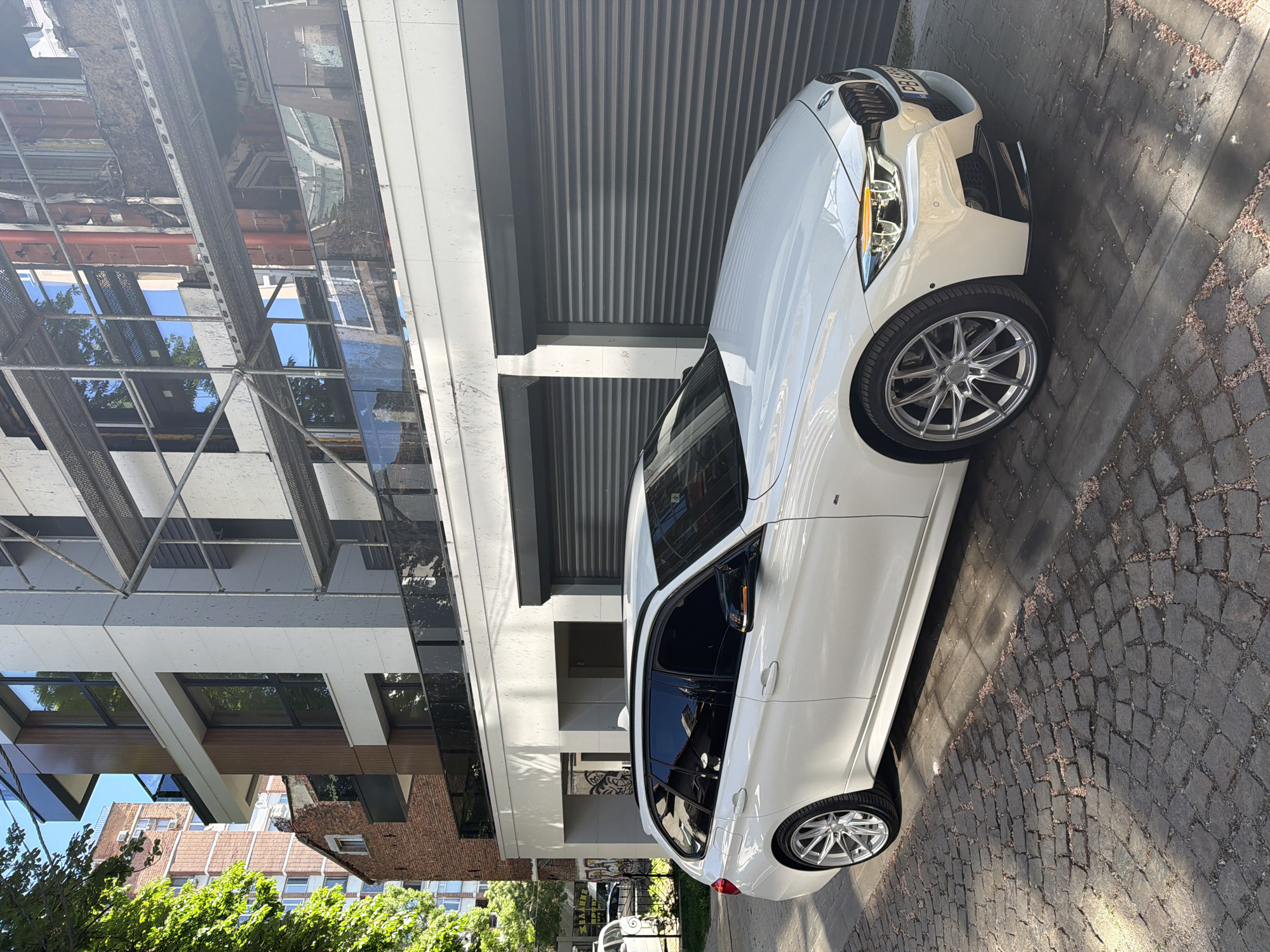 A white BMW sedan is parked on a cobblestone street in front of a modern building in Sofia, Bulgaria. The building features a mix of glass, white concrete, and steel elements. Sunlight illuminates the scene, casting shadows on the pavement. The car is angled towards the viewer, showcasing its front and side profile. Other buildings and greenery are visible in the background.