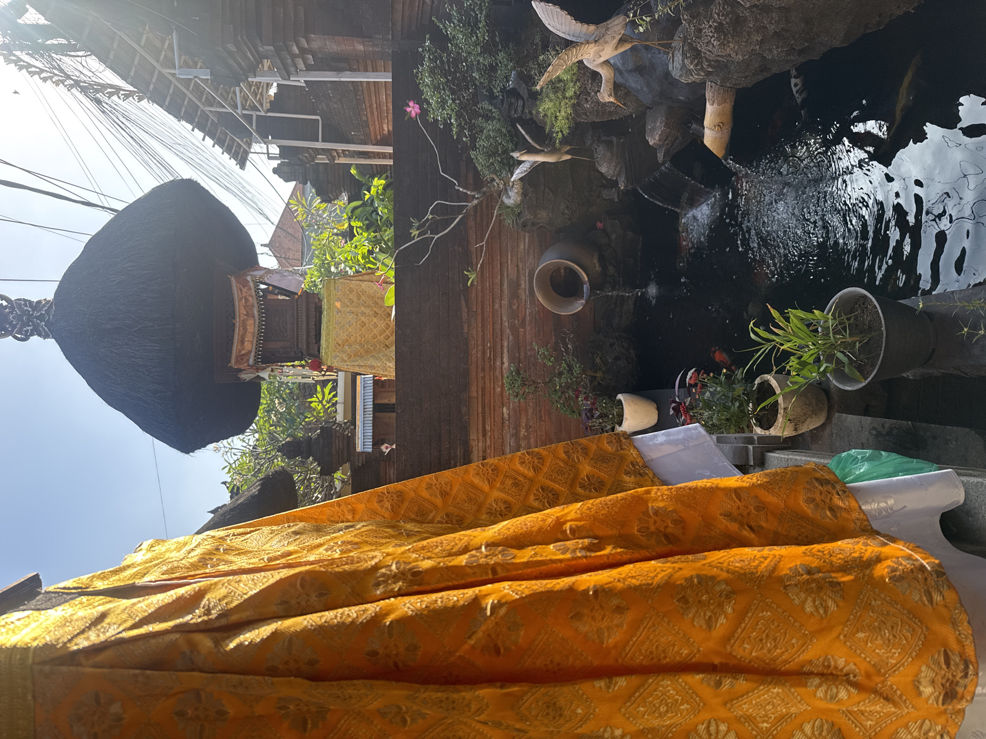In North Kuta, Indonesia, a vibrant outdoor scene unfolds with elements of Balinese architecture and nature. In the foreground, a large, draped piece of orange fabric with a gold patterned print hangs on the left side of the frame. Behind this, a tiered structure with a thatched roof stands under a clear blue sky, adorned with ornate carvings and decorative hangings. Below this structure, a serene pond is visible, populated with koi fish. A small waterfall flows into the pond from a large ceramic pot. The pond area is landscaped with rocks, various plants, and decorative wooden bird statues. The background features brick walls and the roofs of traditional Indonesian buildings, intertwined with a dense network of electrical wires. The sun casts bright light, creating distinct shadows and highlights throughout the scene.