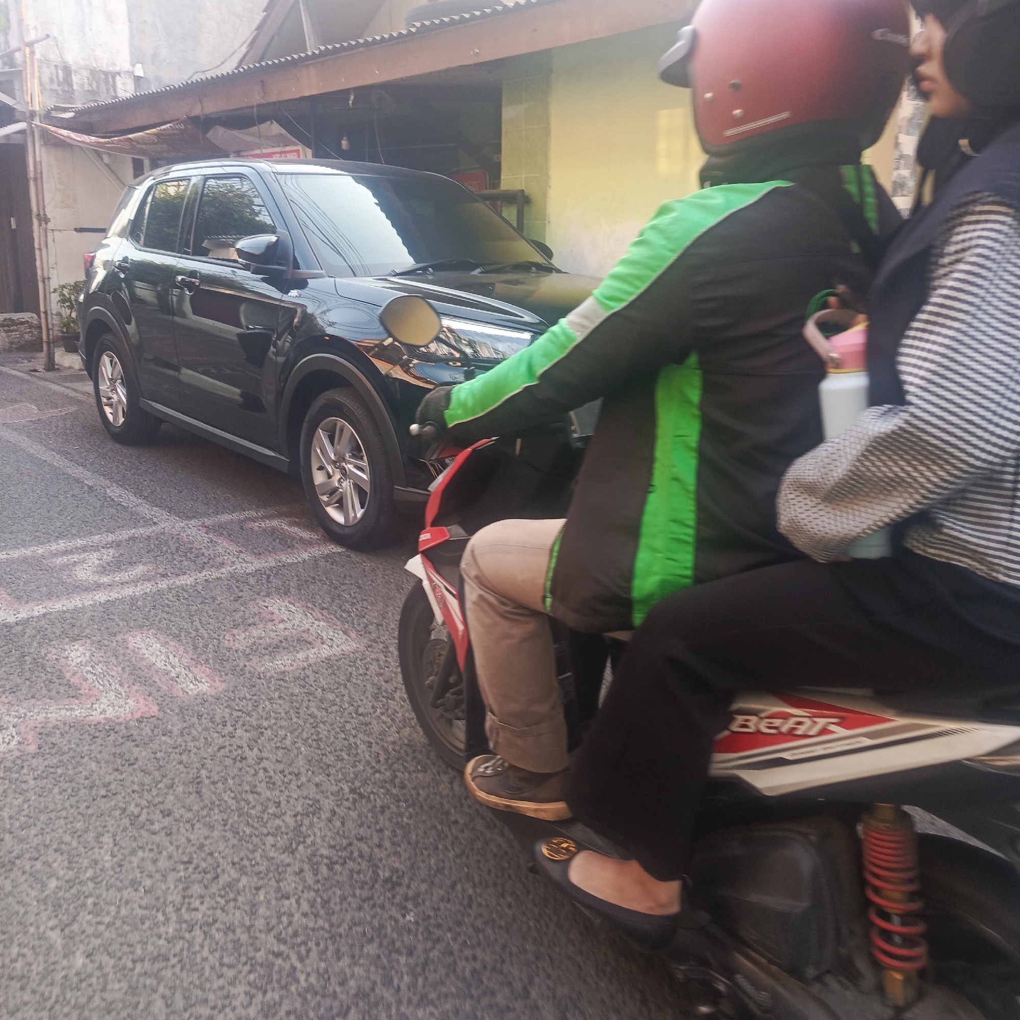 A black SUV is parked on the left side of a narrow city street. In the foreground, two people are riding a motorcycle, with the driver wearing a red helmet and a black and green jacket, and the passenger wearing a dark blue hijab and a striped jacket. The motorcycle is facing away from the camera. The street has painted markings on the asphalt. The background shows buildings with corrugated metal roofs and some vegetation. This scene is typical of a street in South Jakarta, Indonesia.