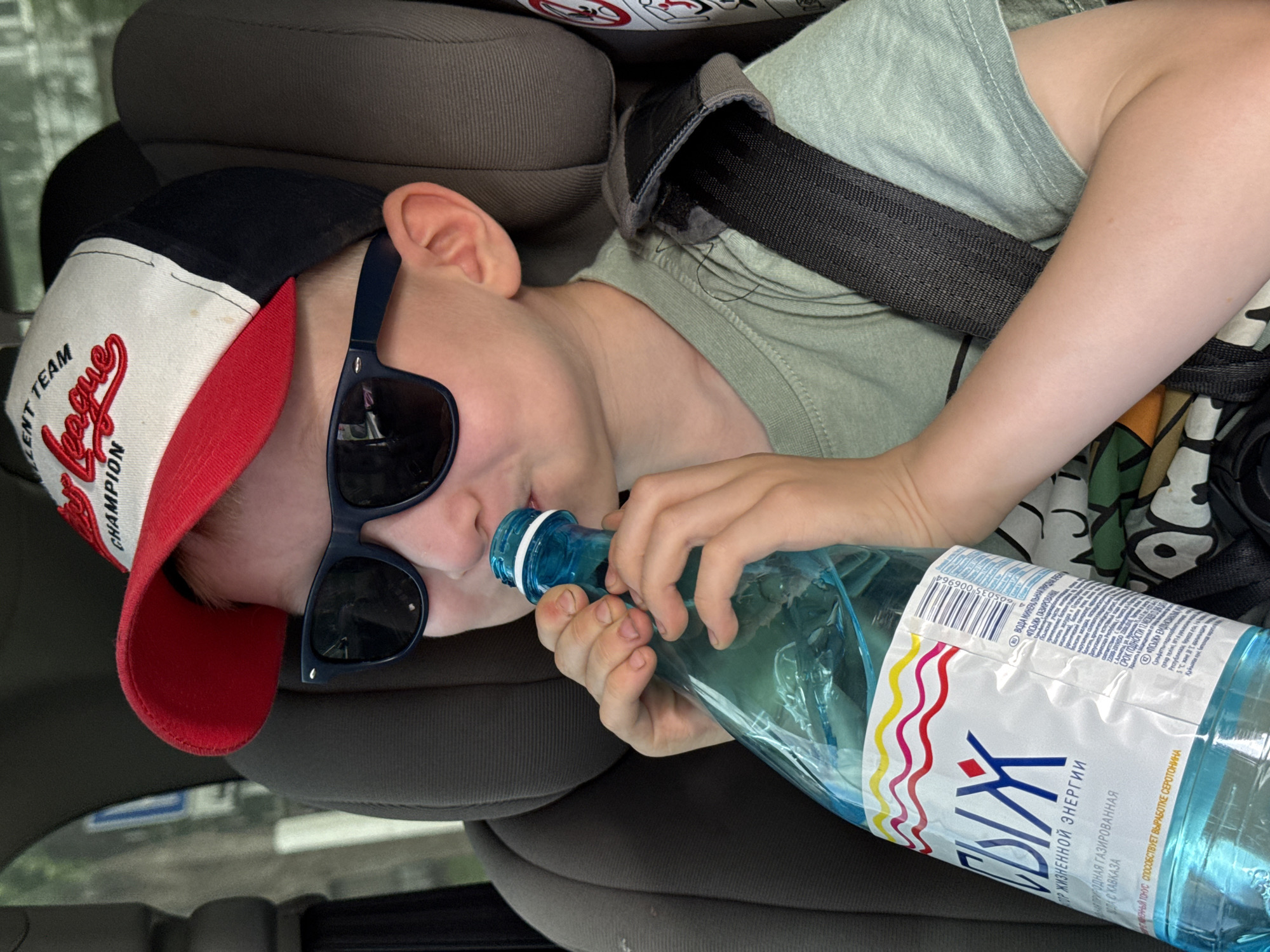 The media file shows a young boy lying down in a car seat, wearing a baseball cap, sunglasses, and a light green t-shirt. He is holding a blue plastic bottle of sparkling mineral water and appears to be drinking from it. The cap is red and white with the words "LENT TEAM CHAMPION" visible. The bottle has a label with colorful wavy lines and text in Russian. The overall scene suggests a casual setting, possibly during a car journey on a bright day. The boy's pose and the presence of a car seat indicate he is a passenger. There is text on the bottle label, including brand names and information about the water's origin. The location context provided is Chaikovsky, Russia.