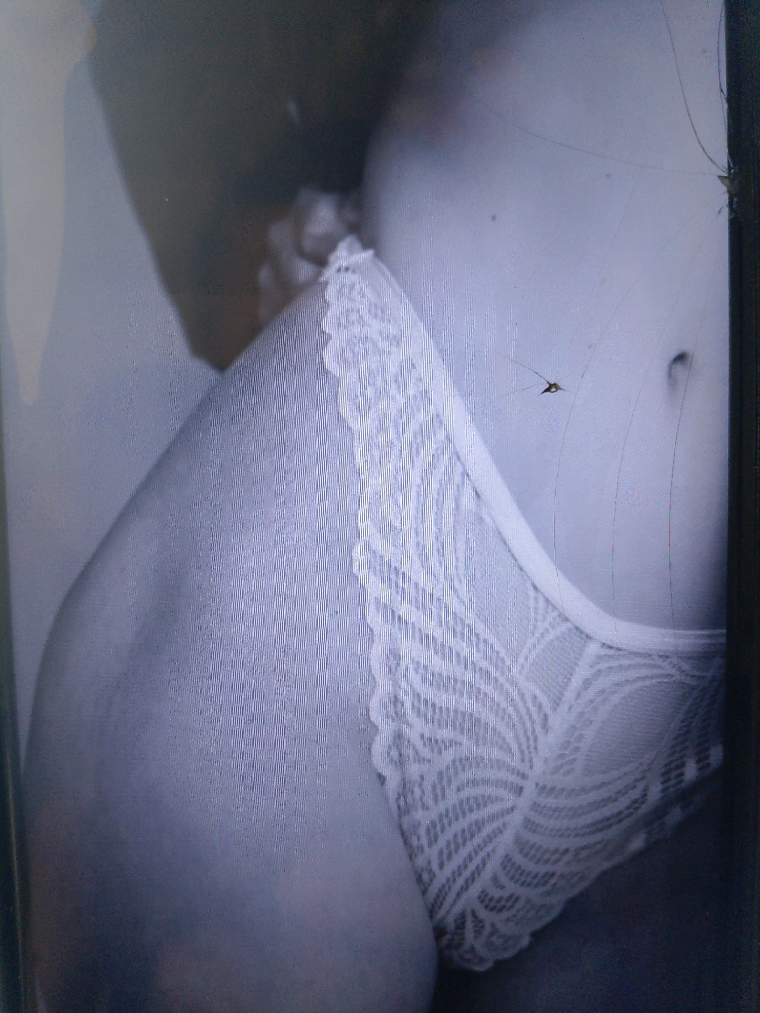 The image displays a close-up view of a person's torso wearing white lace underwear. The lighting is dim, and the image appears to be taken with a phone camera due to the visible screen glare and a cracked screen overlay. The background is dark and indistinct. The body part visible includes the abdomen and hip area. The focus is on the texture and detail of the lace. There is no indication of the location being Dobra Krynytsia, Ukraine, or any other specific geographical location. The scene is static, with no observable actions or interactions.