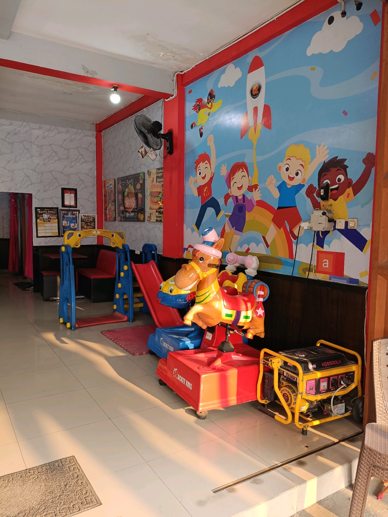 The image depicts an indoor play area, likely a section of a restaurant or cafe, located in Ngemplak, Indonesia. The space features a vibrant mural on the wall depicting cartoon children with a rocket. In the foreground, a red and yellow coin-operated kiddie ride resembling a horse is positioned. To the left of the kiddie ride is a small blue and yellow children's playset with a red slide. Further left, a row of dark seating with red cushions is visible, set against a textured light gray wall. On the wall, several framed posters and notices are displayed. A ceiling fan is mounted on the left wall. In the right foreground, a yellow portable generator is situated. The floor is tiled in a light cream color, with long shadows cast across it from the sunlight entering from the right. The overall impression is one of a child-friendly environment within a commercial establishment.