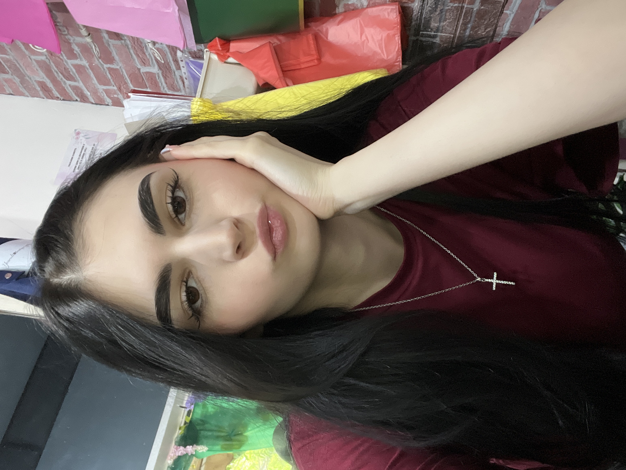 A young woman with long, black hair is posing for a photograph. She is wearing a maroon top and a silver necklace with a cross pendant. Her left hand is resting on her face. The background includes a brick wall and various colored objects, potentially paper or plastic bags. The image does not provide definitive information about Burgas, Bulgaria.