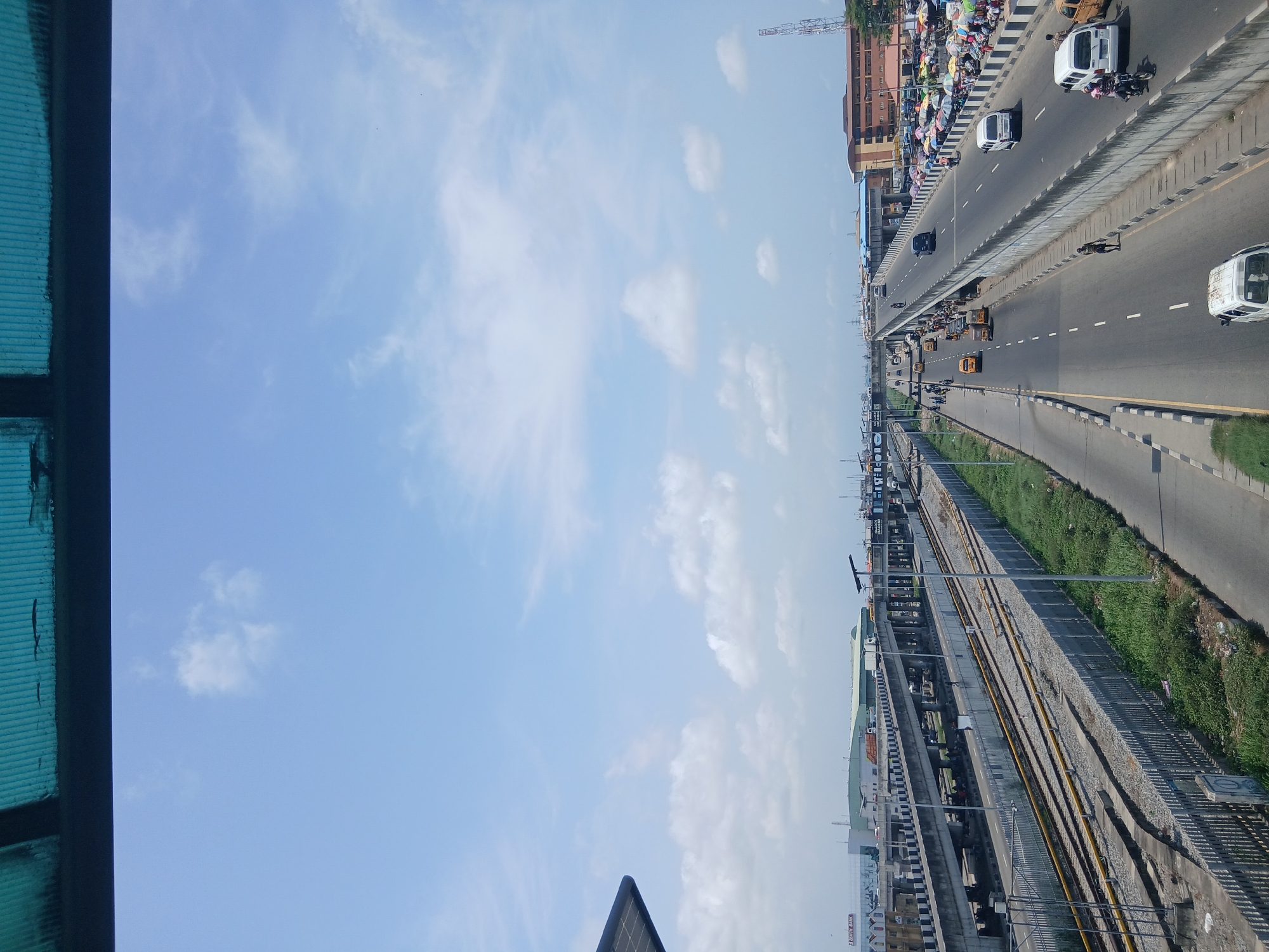 An elevated view of Alaiyabiagba, Nigeria. A multi-lane highway carries light traffic. Adjacent to the highway, elevated train tracks and a station are present. Street vendors and pedestrians crowd the edge of the highway. The sky is mostly clear with scattered clouds. The view is from a higher vantage point, possibly another elevated structure.