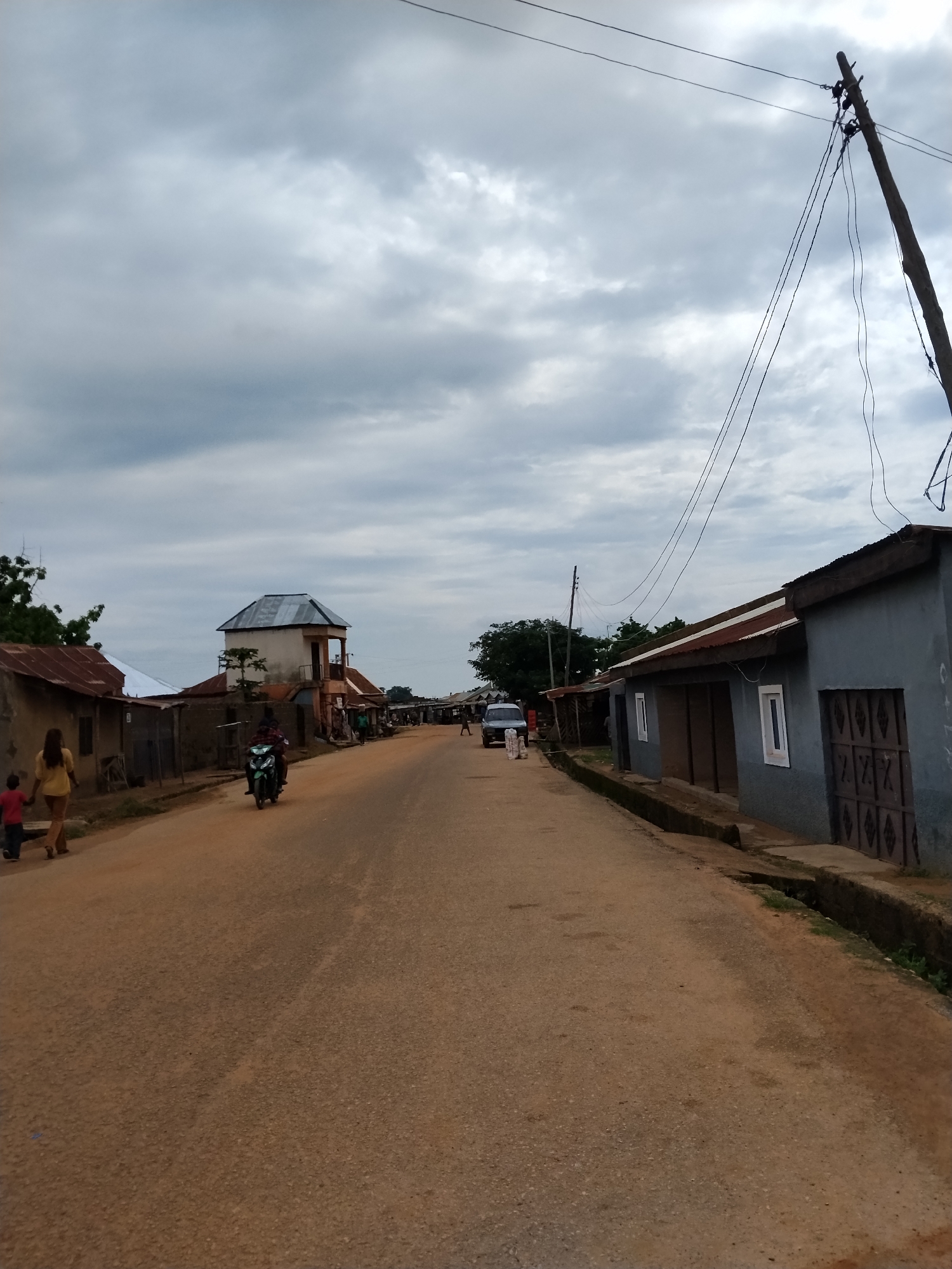 This is a medium-shot, eye-level, outdoor photograph taken on a cloudy day, capturing a street scene in Mangu Town, Nigeria. The unpaved dirt road runs through the center of the image, lined with buildings on both sides.
On the left side of the road, a woman in a yellow top and brown trousers is walking with a child in a red shirt. Further down the road on the left, a green motorcycle with two riders is visible. Behind the motorcycle, several low-lying buildings with corrugated metal roofs can be seen. One building stands out with a more elaborate design, featuring a square tower with a metal roof and balconies.
On the right side of the road, a row of light blue buildings with corrugated metal roofs stretches into the distance. These buildings have a simple design with a few windows and decorative metal doors. A concrete drainage channel runs along the edge of the road in front of these buildings.
In the background, more buildings and trees are visible, with a utility pole and power lines stretching across the sky. A silver SUV is parked on the left side of the road in the mid-ground. The overall impression is that of a somewhat underdeveloped urban or semi-urban environment.