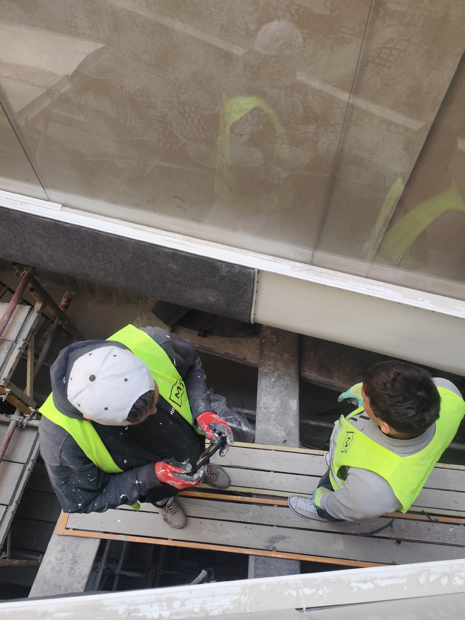 This high-angle image depicts two construction workers on a scaffolding platform, engaged in work on a building structure. Above them, a large glass panel heavily reflects light and shows numerous distinct boot or shoe prints, alongside reflections of the workers themselves and the scaffolding.
The worker on the left is viewed from directly above, wearing a white baseball cap reversed, a dark long-sleeved top splattered with white paint, and red work gloves. His bright yellow high-visibility vest bears a logo featuring "MJ" and the word "JUST." He is in a crouched or bent position, appearing to be examining a dark object held in his gloved hands, possibly a mobile phone or a small tool. He wears light brown work shoes.
The second worker, positioned to the right, has dark hair and is wearing a grey long-sleeved shirt with light blue/green work gloves. His bright yellow high-visibility vest also displays an "MJ" logo, along with the text "KV." He stands on the wooden planks of the scaffolding platform, sporting white canvas sneakers, and appears to be looking downwards or towards the first worker.
The setting includes a scaffolding structure of metal poles and wooden planks beneath and around the workers. Above them, a dark grey, textured building panel is visible, bordering a white horizontal beam that sits beneath the reflective glass surface. The numerous shoe patterns on the glass are a prominent detail, suggesting dust, dirt, or foot traffic on the surface, which might be a skylight or a large window. The reflections of the workers' yellow vests are also clearly visible on this glass. The scene is illuminated by natural light, indicating it is daytime.