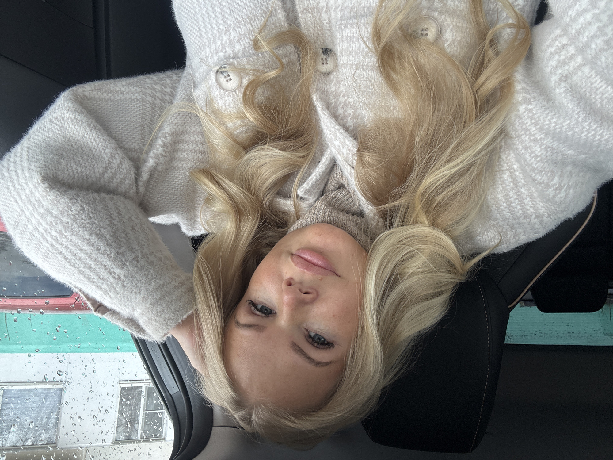 A close-up, upside-down portrait features a young woman with long, wavy blonde hair and light-colored eyes, looking directly at the camera with a neutral expression. Her head is positioned towards the bottom of the frame, resting against a dark surface, likely a car seat headrest. She wears a light grey or off-white textured coat or cardigan with large white buttons, over a beige ribbed turtleneck sweater.
Her blonde hair is spread out around her head and shoulders. To the left, a car window heavily covered in raindrops is visible, suggesting it's raining or has recently rained. Through the distorted view of the wet window, an inverted outdoor scene is faintly visible, showing a building with white walls and dark-framed windows, and above it, horizontal bands of red and turquoise/green, possibly part of another structure or vehicle. The lighting is soft and diffused, indicative of an overcast day. No text is visible in the image.