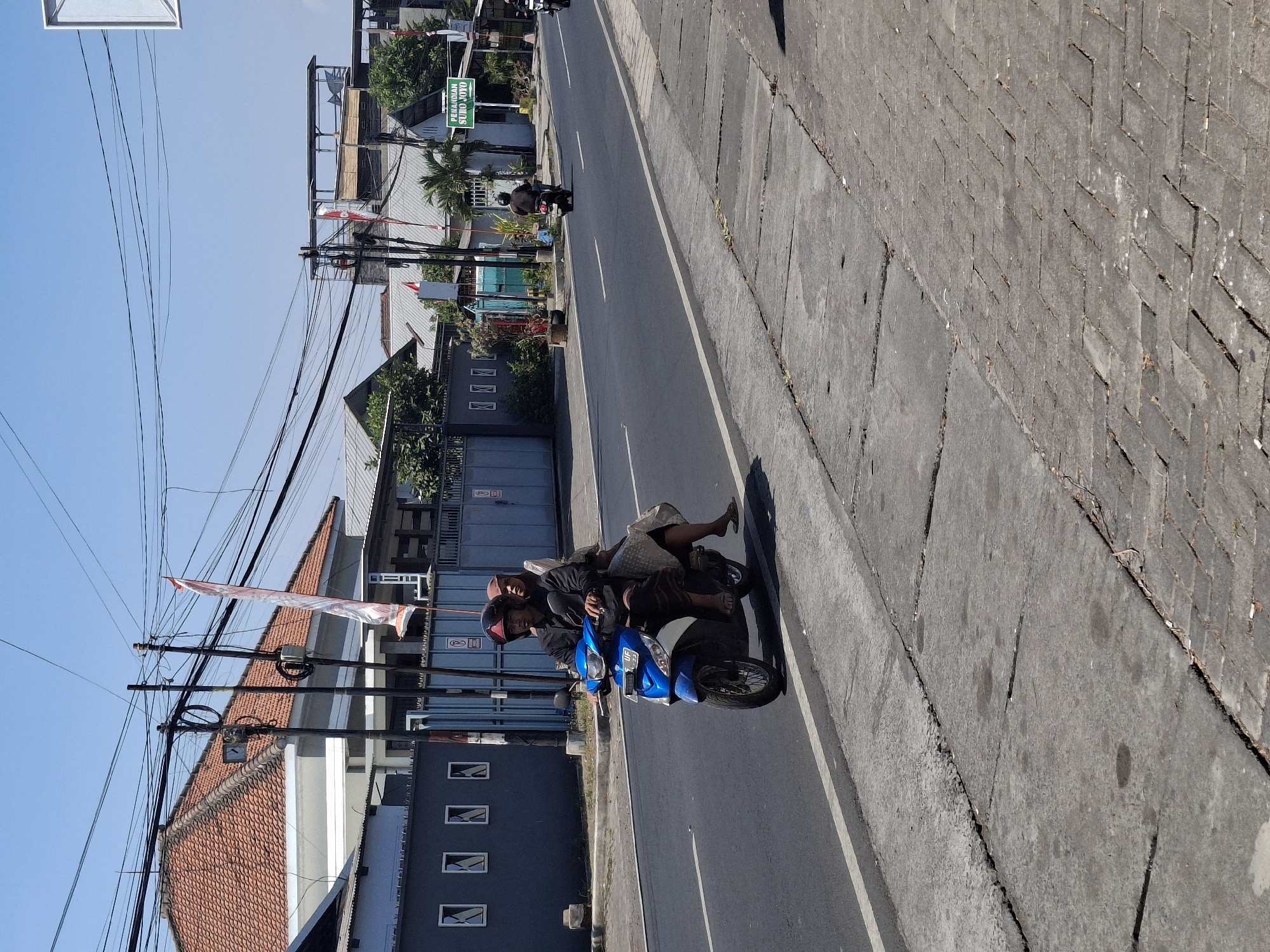 In Lumajang, Indonesia, two individuals are riding on a blue and white scooter. The person in front, wearing a dark long-sleeved shirt and a red helmet, is driving. The person behind, wearing a light-colored head covering and a patterned skirt, is a passenger. They are traveling on a paved road with a gray concrete sidewalk. Several buildings with orange tiled roofs and dark gray walls are visible on the left side of the road. Overhead, a network of electrical wires crisscrosses against a clear blue sky. In the distance, a green sign with white lettering is mounted on a pole. Another scooter is seen further down the road.