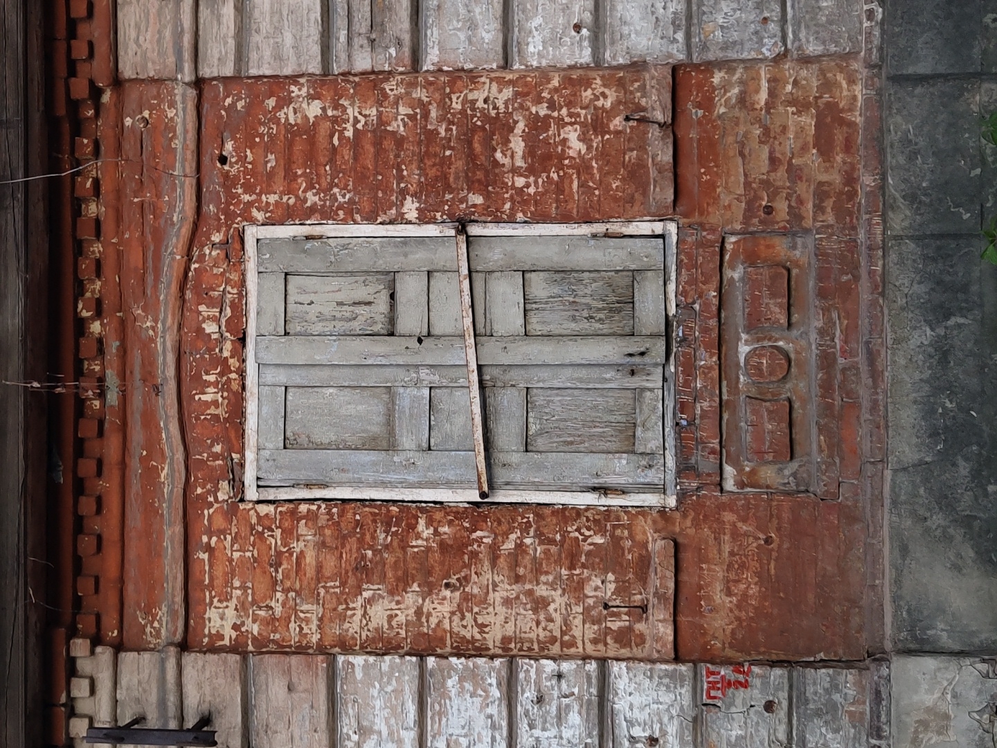 A closed, weathered wooden window with a white frame is centered in the image, mounted on a wall of decaying brown and orange wood. The wood is textured with flaking paint and a rustic appearance. To the left of the window, a vertical wooden structure with a corrugated pattern is visible, while to the right is a panel with a series of rectangular and circular shapes embossed into it. The bottom of the frame is bordered by weathered white wooden planks. The overall impression is one of age and disrepair. The location is not determinable from the image.