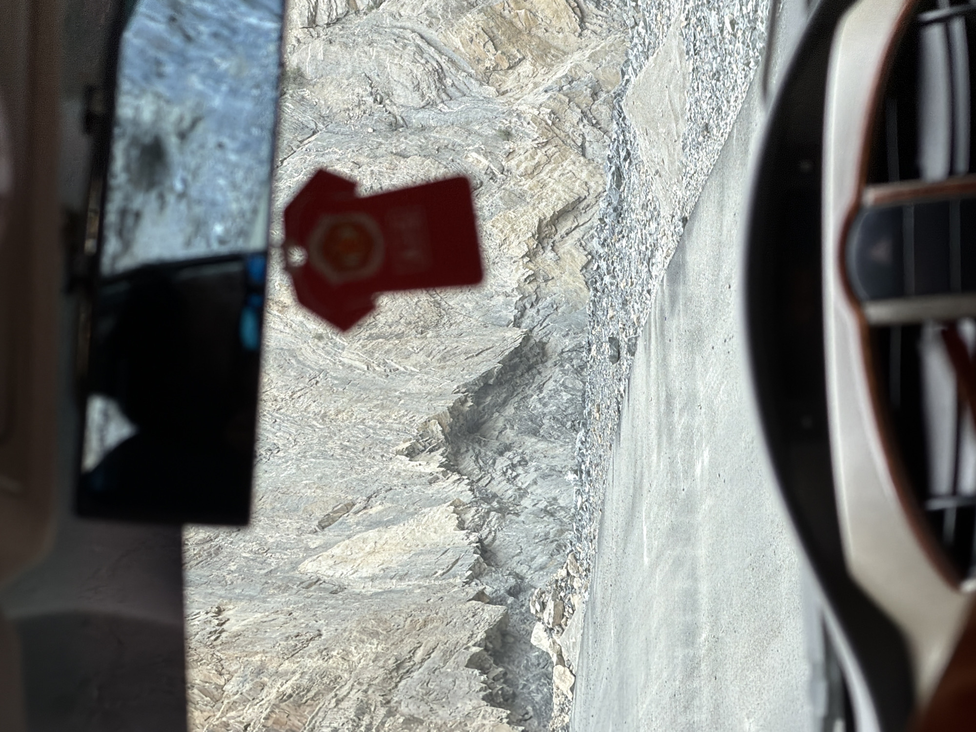 A first-person perspective from inside a vehicle shows a rough, gray asphalt road winding through a mountainous landscape. The road is flanked by a steep, rocky cliff face composed of layered gray and tan stone. Loose rocks and scree are scattered along the base of the cliff and on the shoulder of the road. In the foreground, the blurred dashboard of the vehicle is visible, including a portion of the steering wheel and center console. Hanging from the rearview mirror is a small, red t-shirt-shaped air freshener with a circular emblem. The scene suggests travel through a remote, rugged, and possibly hazardous terrain. The image location is Stongde, India.